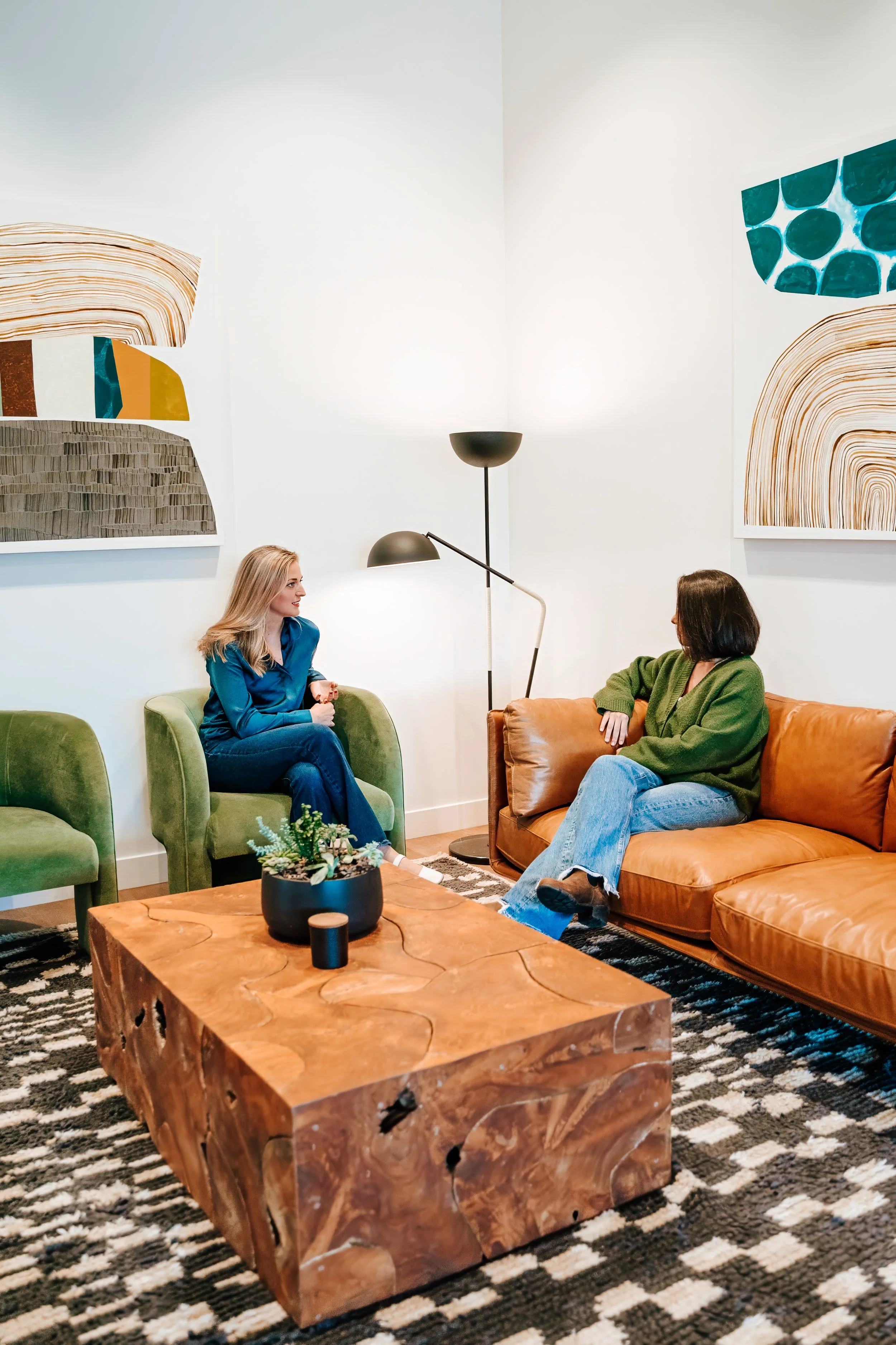 Two women sitting on sofas in a modern living room, conversation in progress, with abstract artwork on the walls and a wooden coffee table with a potted plant in the center.