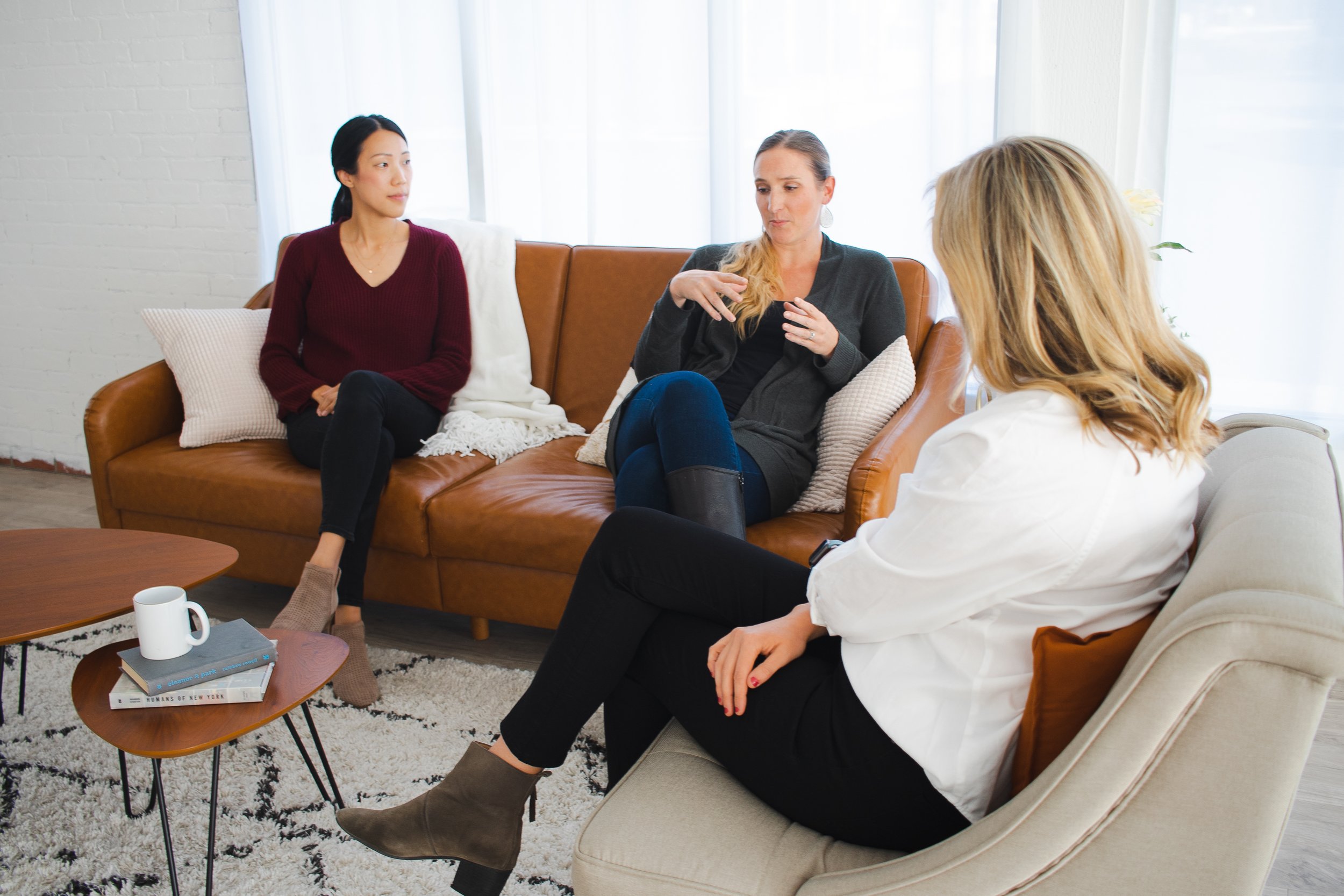 Three women sitting on sofas in a brightly lit room, engaged in conversation, with a coffee table holding books, a mug, and a book on the floor.
