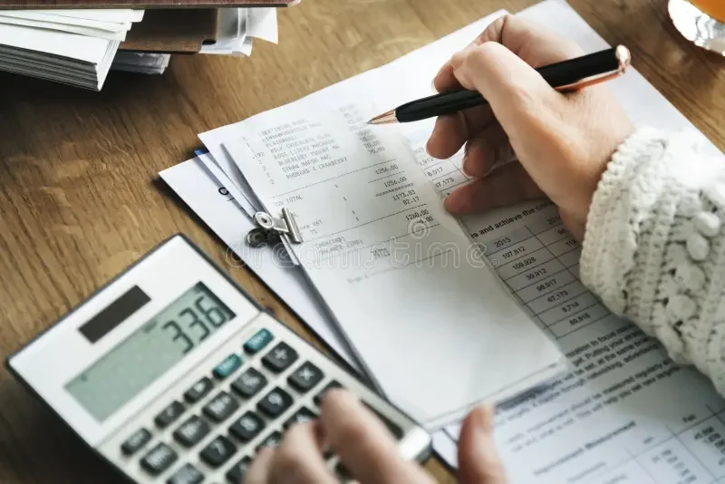 A person with a sweater writing with a pen on some papers, a calculator showing 336, and stacks of documents on a wooden desk.
