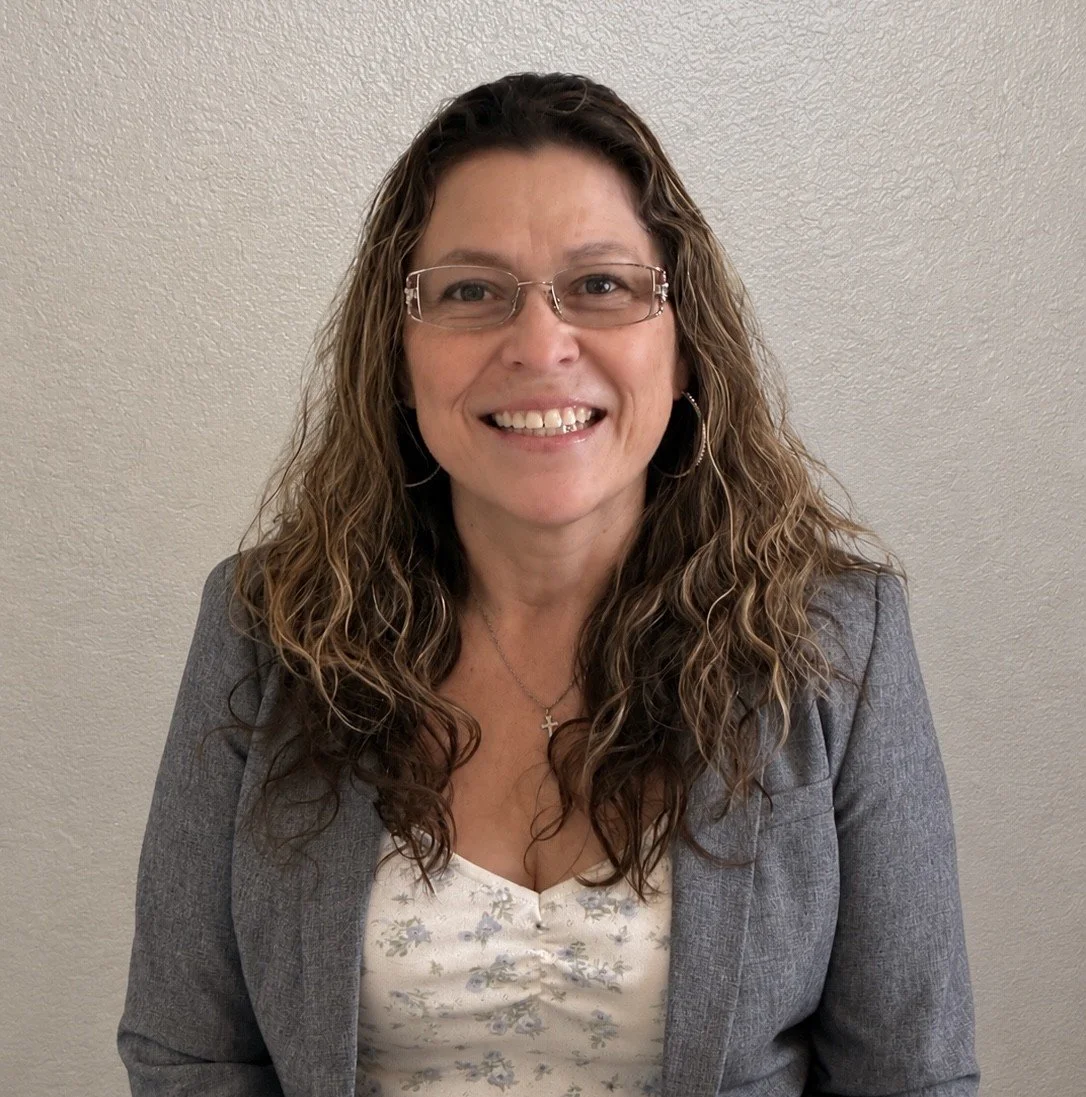 A woman with curly brown hair wearing glasses, a gray blazer, and a floral top, smiling in front of a plain wall.