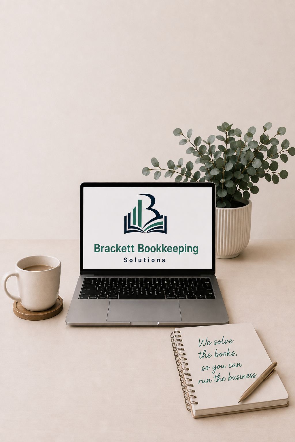 A desk setup with a laptop displaying the logo of Brackett Bookkeeping Solutions, a cup of coffee on a wooden coaster, a spiral notebook with a handwritten note saying 'We solve the books, so you can run the business,' a pen, and a potted eucalyptus plant on a beige surface against a plain beige background.
