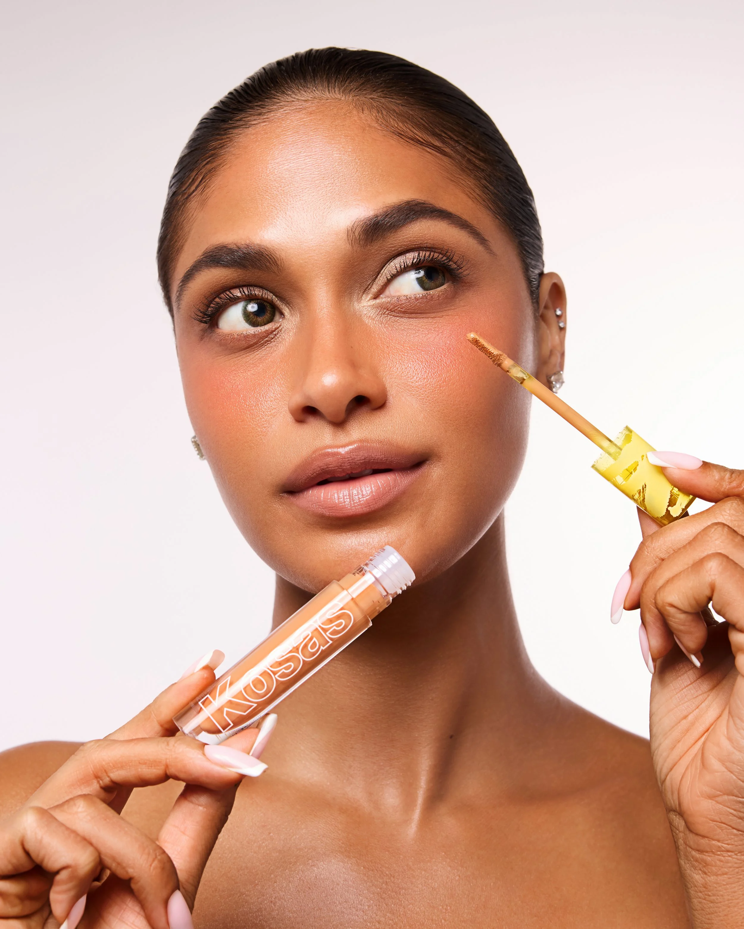 A woman with smooth, tan skin and brown hair slicked back applies a golden makeup product to her face with a small brush. She holds a tube labeled 'GLOSSY' and 'KISS' in one hand, while her other hand supports her face. Her nails are manicured with a