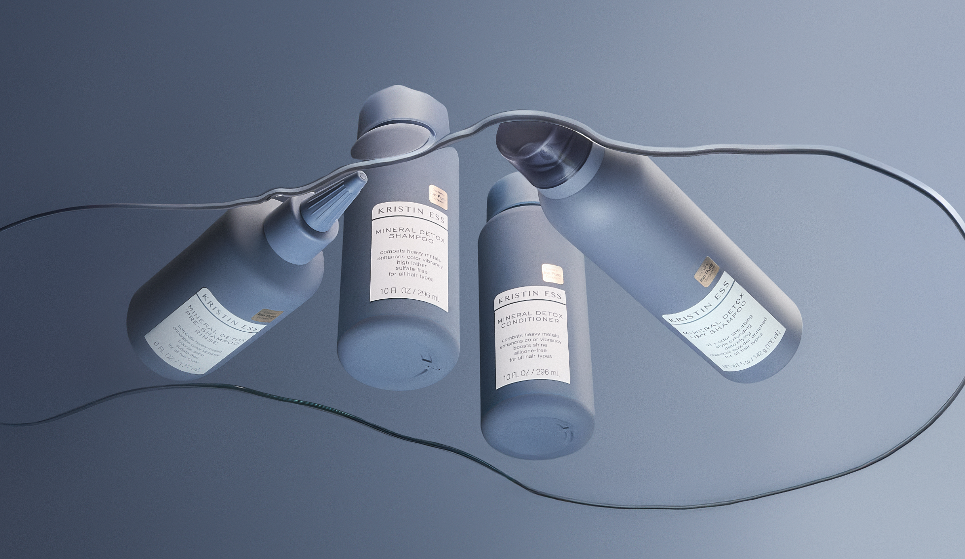 Three bottles of Kristin Ess hair care products, including a mineral detox shampoo, conditioner, and pre-shampoo treatment, hanging on a wire against a gray background.