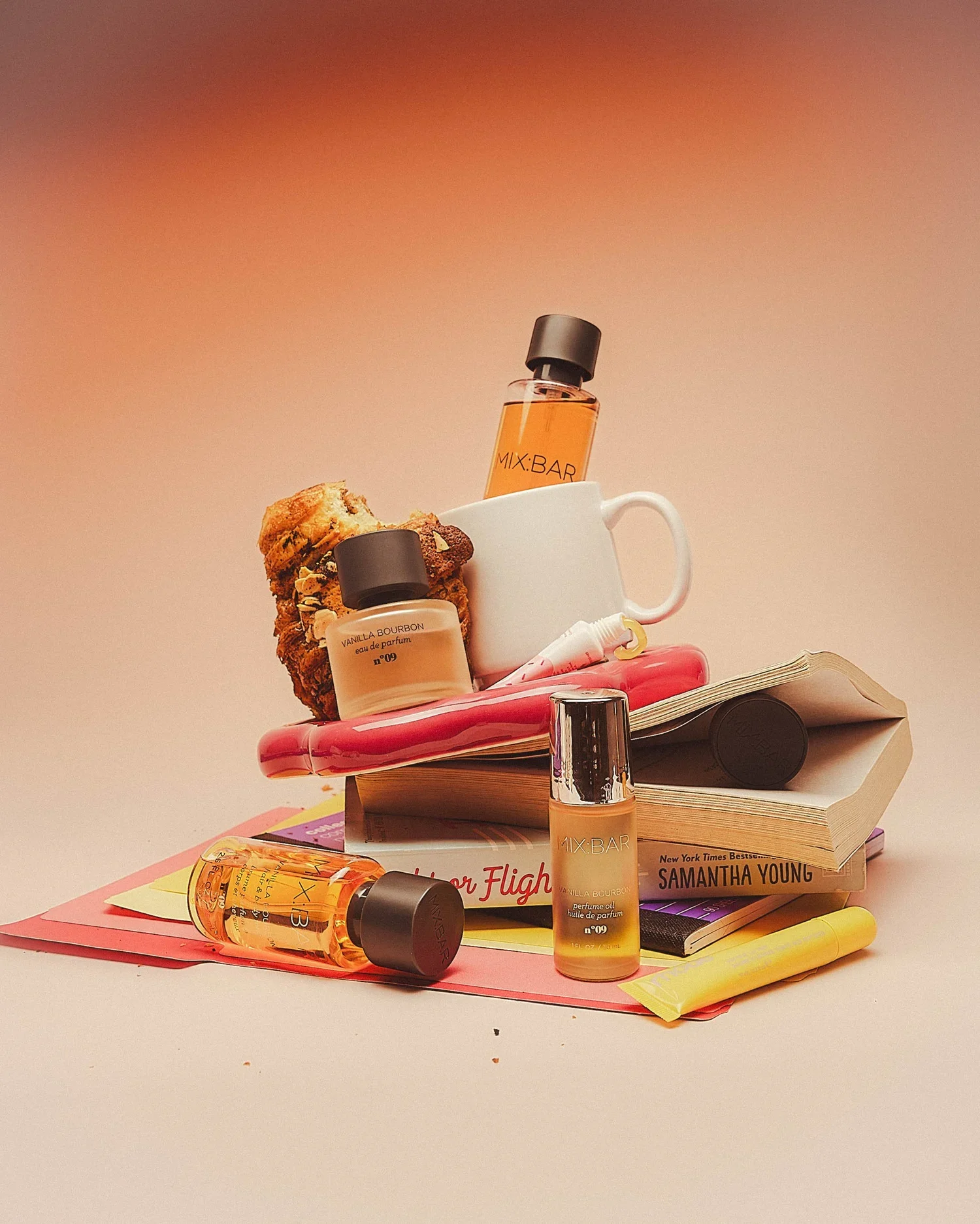 An assortment of beauty and skincare products, books, and snacks arranged on a pink surface against a pink background. Notable items include a vanilla bourbon perfume bottle, a MIX:BAR perfume, a book titled 'Samantha Young', a muffin, and various ot