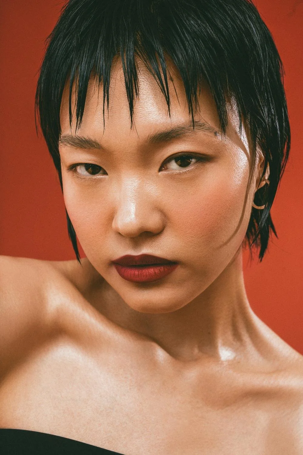 A close-up portrait of a woman with short, black, wet hair, and wearing dark red lipstick, staring intensely at the camera with a red background.