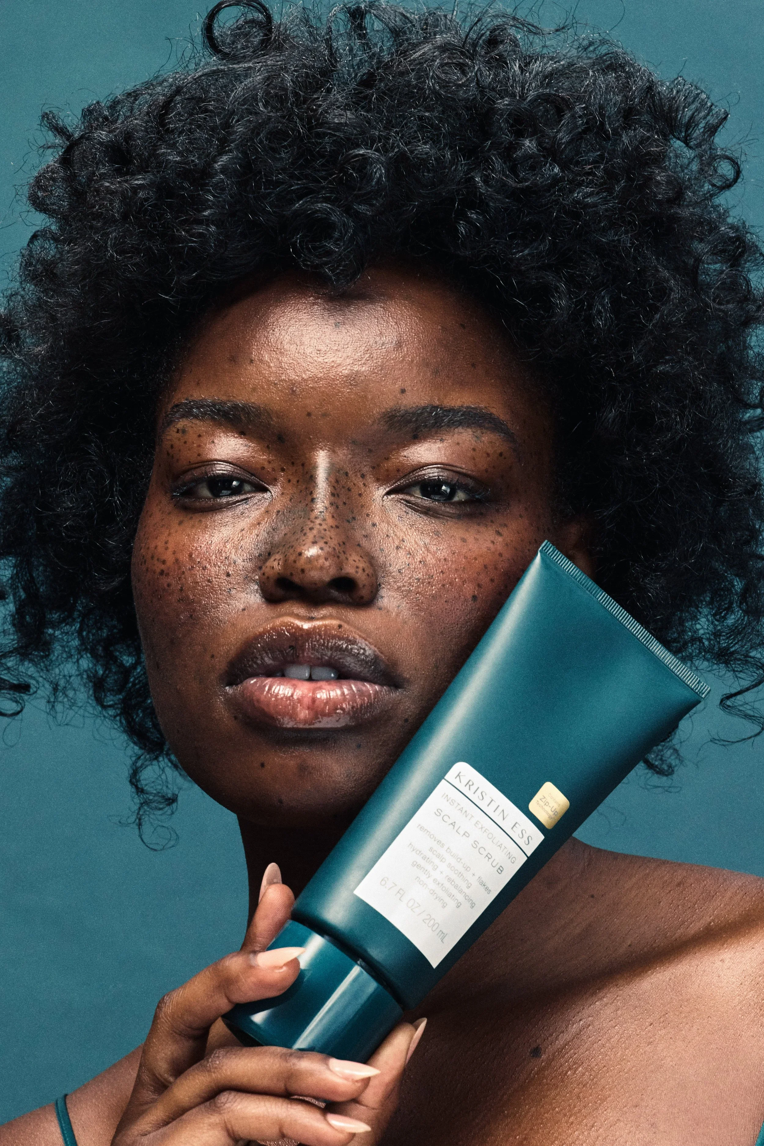 A close-up of a woman with dark skin and natural curly black hair, holding a tube of Kristin Ess scalp scrub. She has freckles across her nose and cheeks and a serene expression, with a teal background behind her.