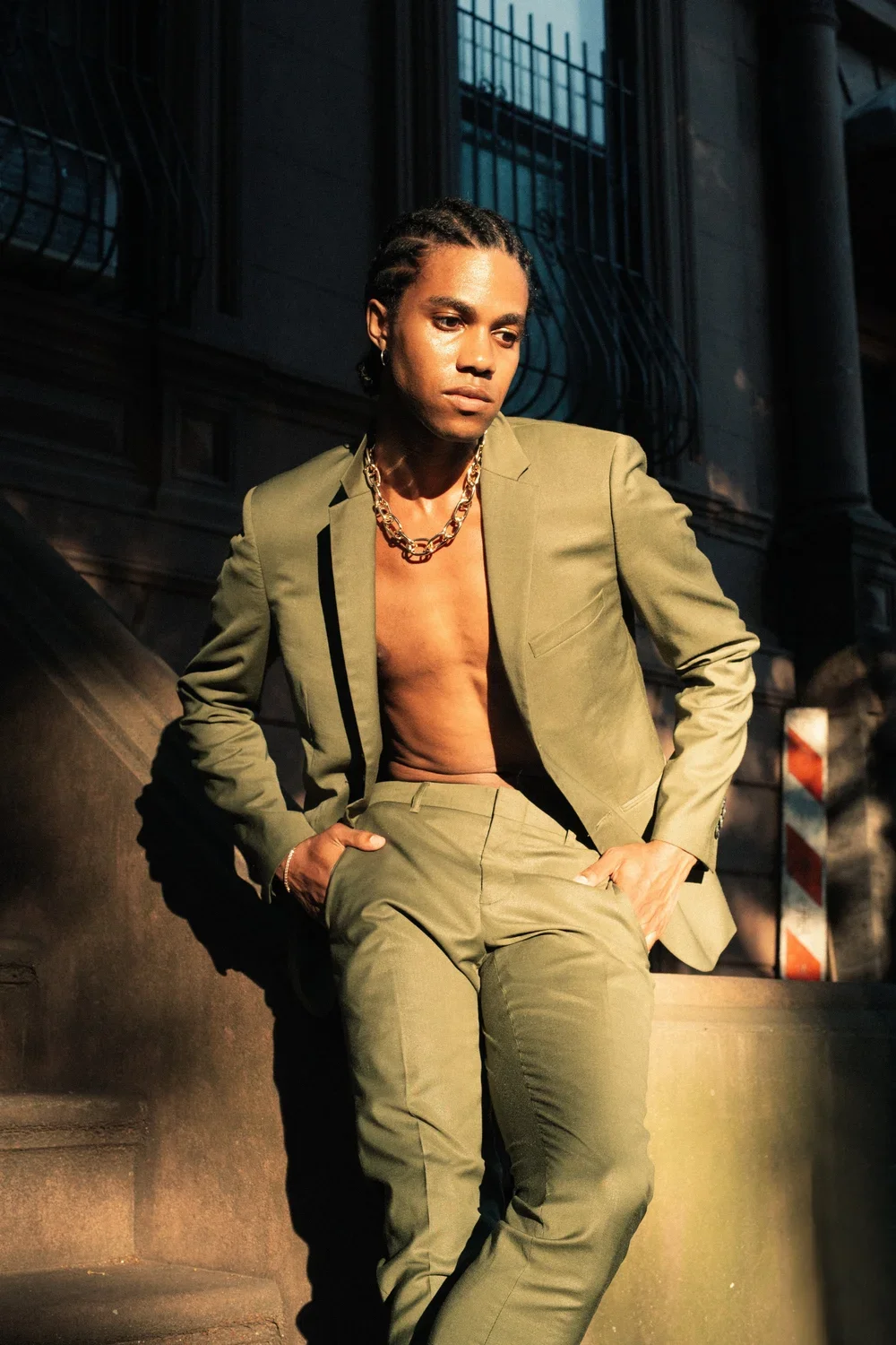 A man with braided hair wearing an olive green suit and a chunky gold chain, standing on a staircase in an urban setting, with one hand in his pocket.