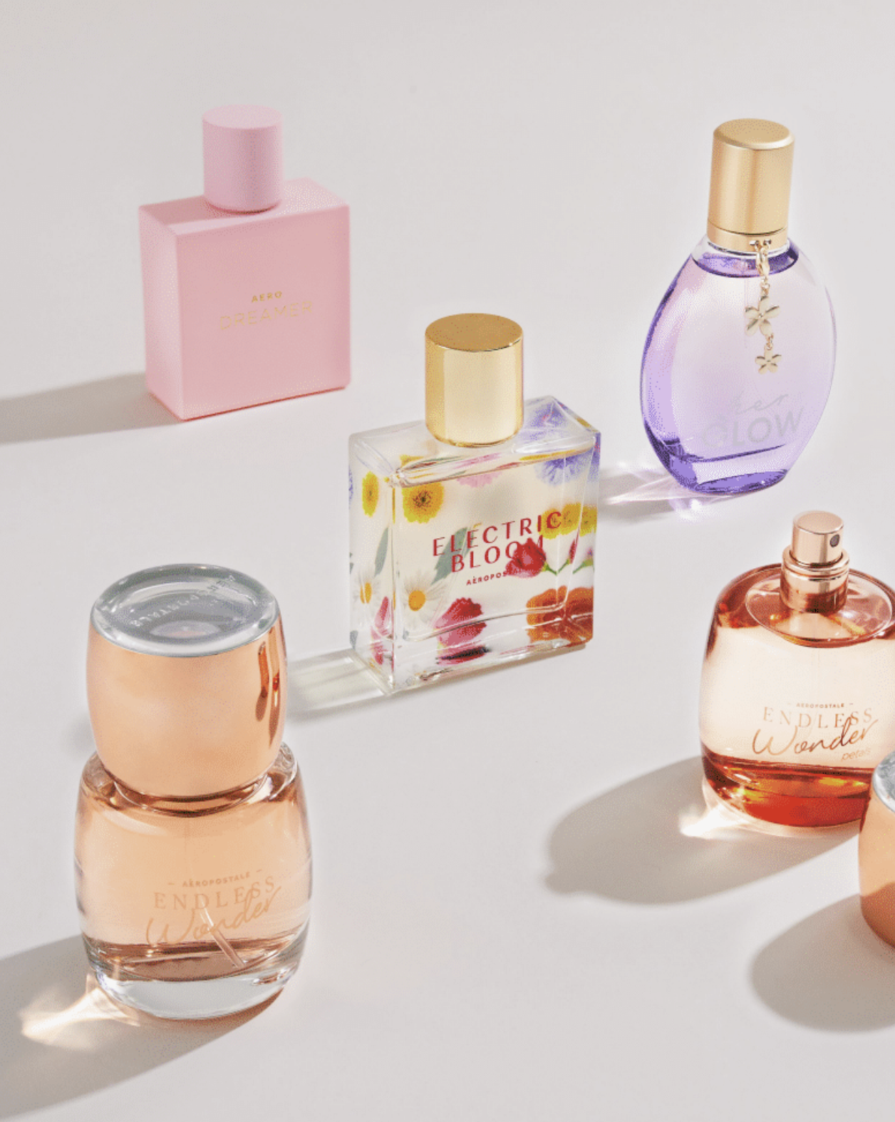 A collection of five perfume bottles on a white surface, including a pink rectangular bottle labeled 'AERØ DREAMER,' a square bottle with floral artwork labeled 'ELECTRIC BLOOM,' a purple oval-shaped bottle with a gold cap and butterfly charm, and two rounded bottles with pinkish liquid, one labeled 'ENDLESS Wonder.'