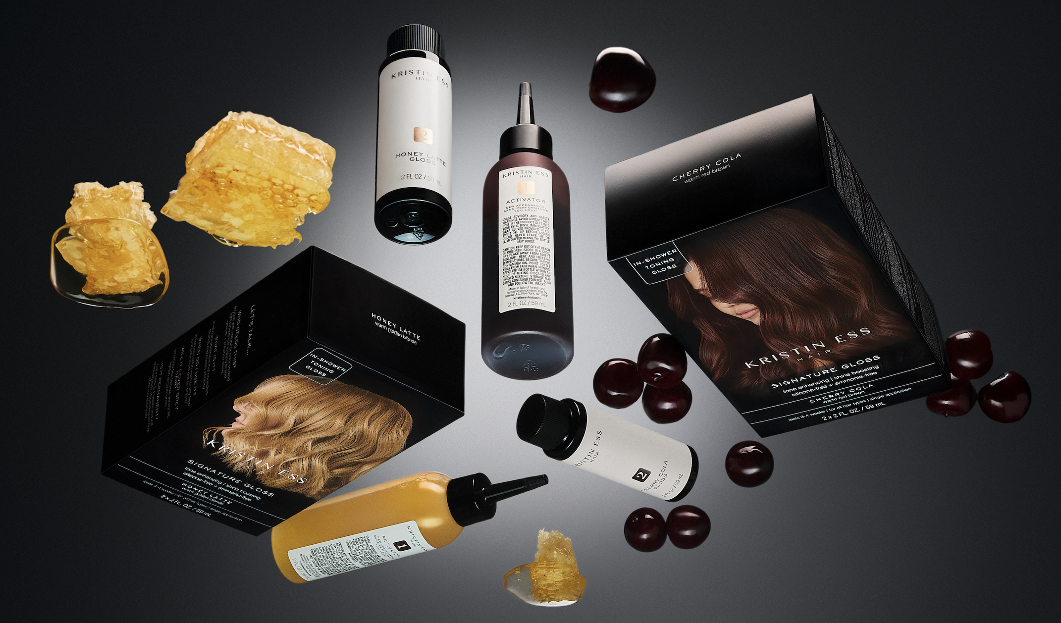Various Kristin Ess hair care products, including shampoo, conditioner, and gloss, along with a box featuring a woman's hair, surrounded by cherries, honeycomb, and a brown cherry on a gray background.