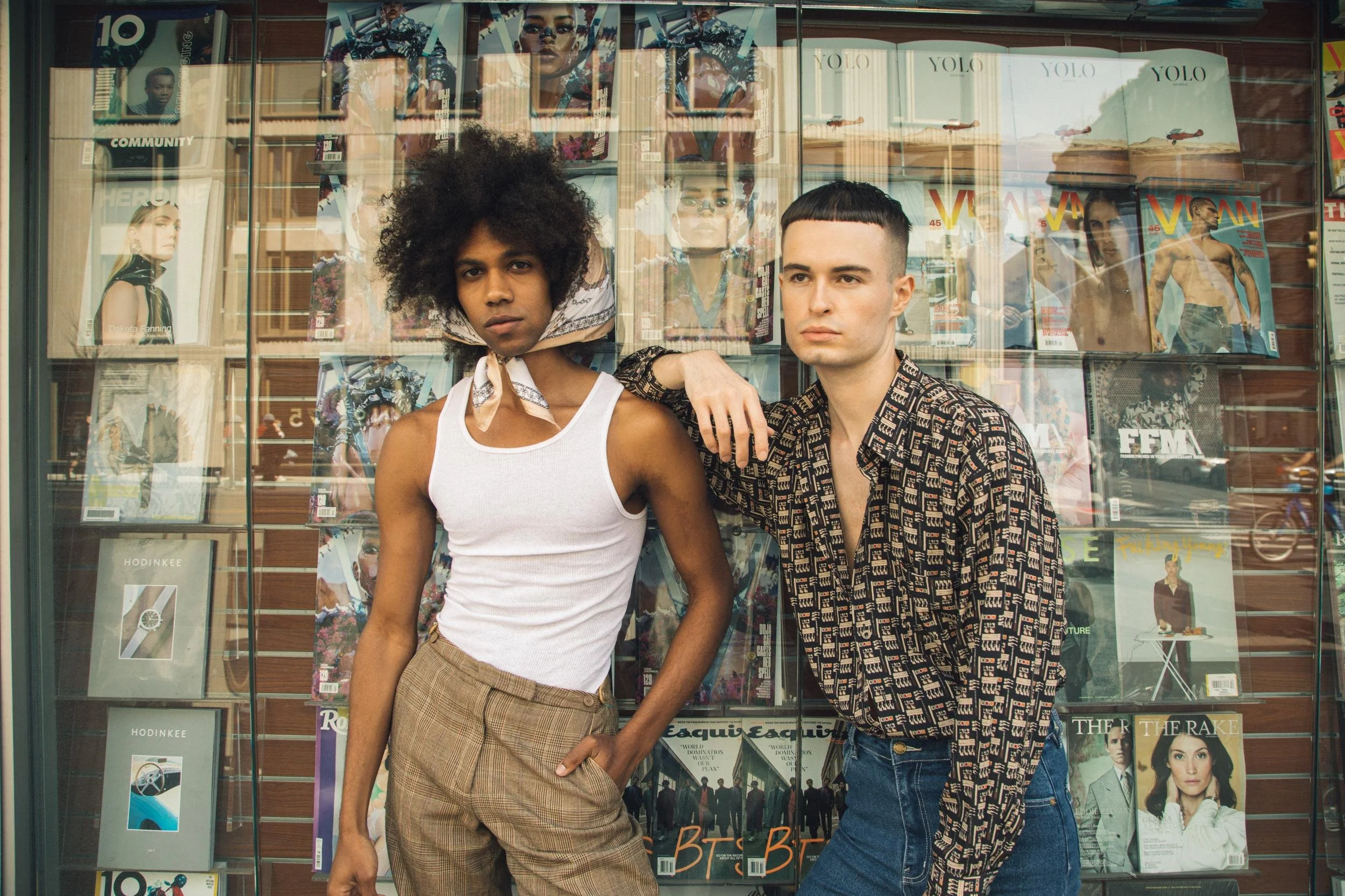 Two young adults standing in front of a magazine display window. The person on the left has dark curly hair, is wearing a white tank top, and beige checkered pants. The person on the right has short straight hair with a fringe, is wearing a patterned