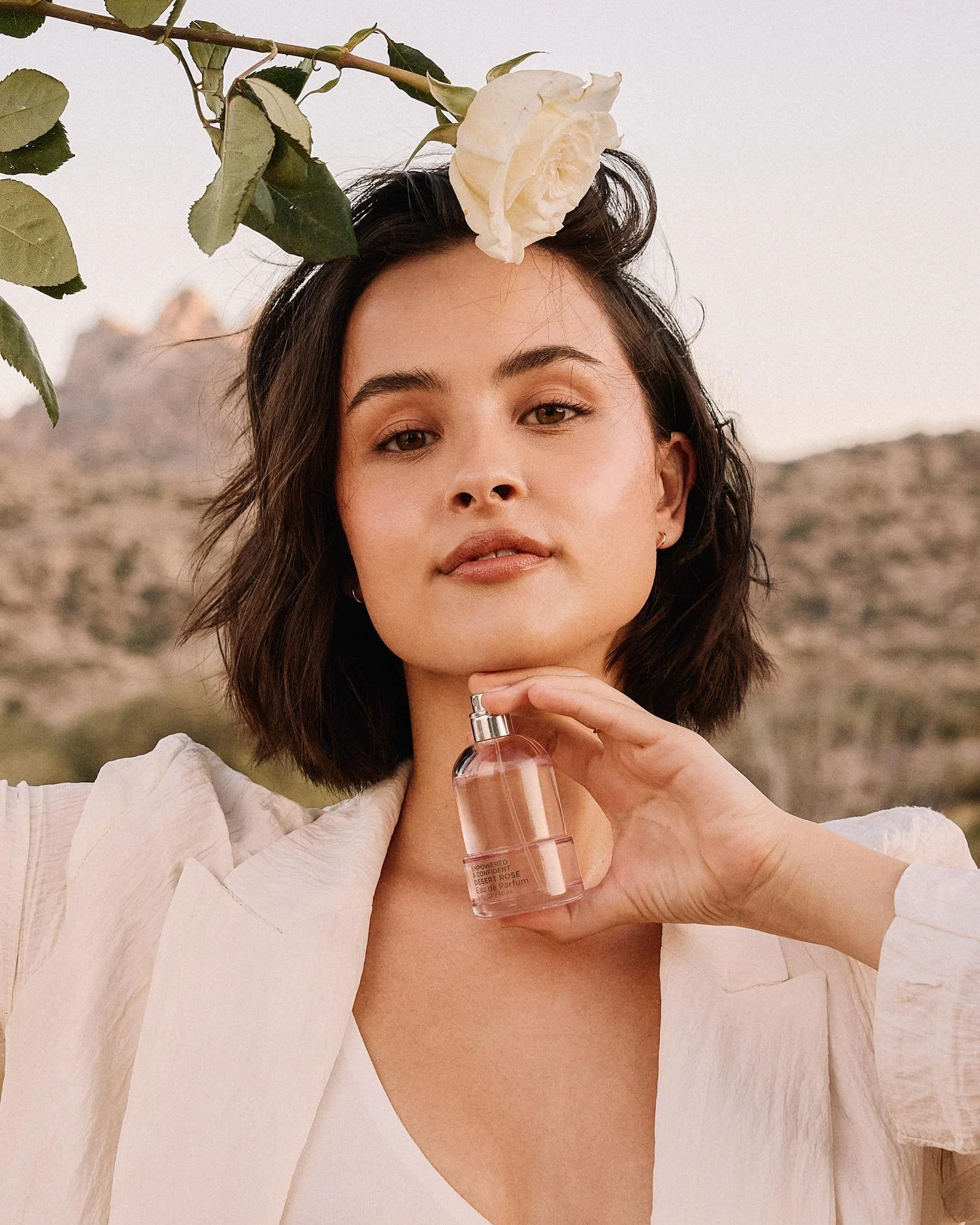 A woman with shoulder-length dark hair holding a small pink perfume bottle near her face, with a white rose nestled in her hair, outdoors with a mountain in the background.