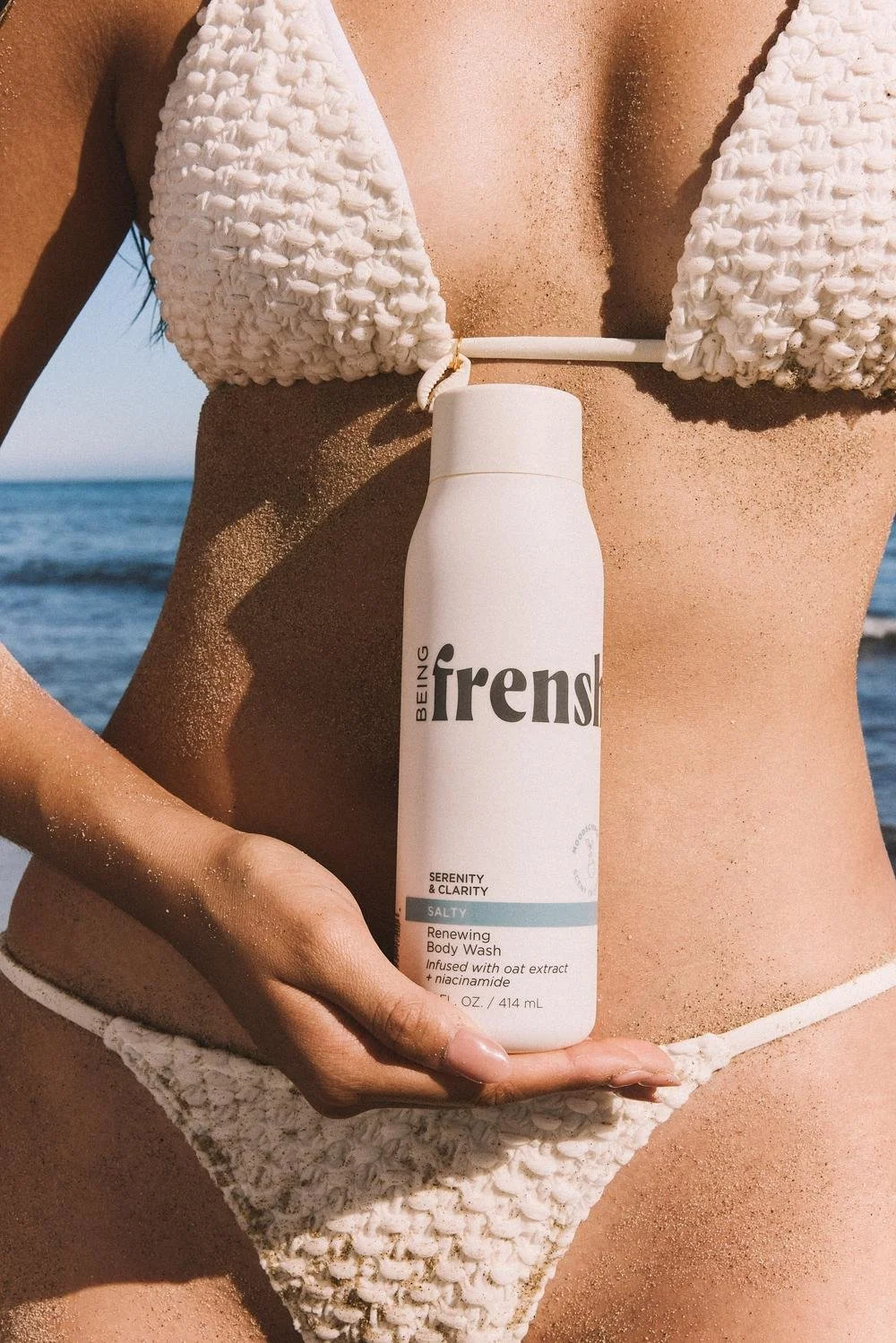 A woman at the beach holding a bottle of Being Trensi body wash in front of her torso. She is wearing a textured white bikini, and the ocean is visible in the background.
