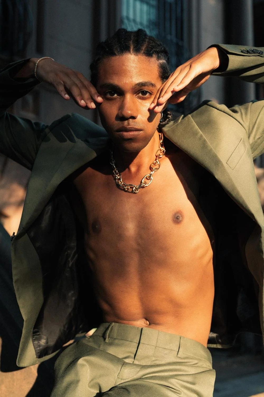 A shirtless young man with braided hair, wearing a green jacket draped over shoulders, a gold chain necklace, and matching green pants, posing outdoors with hands framing face.
