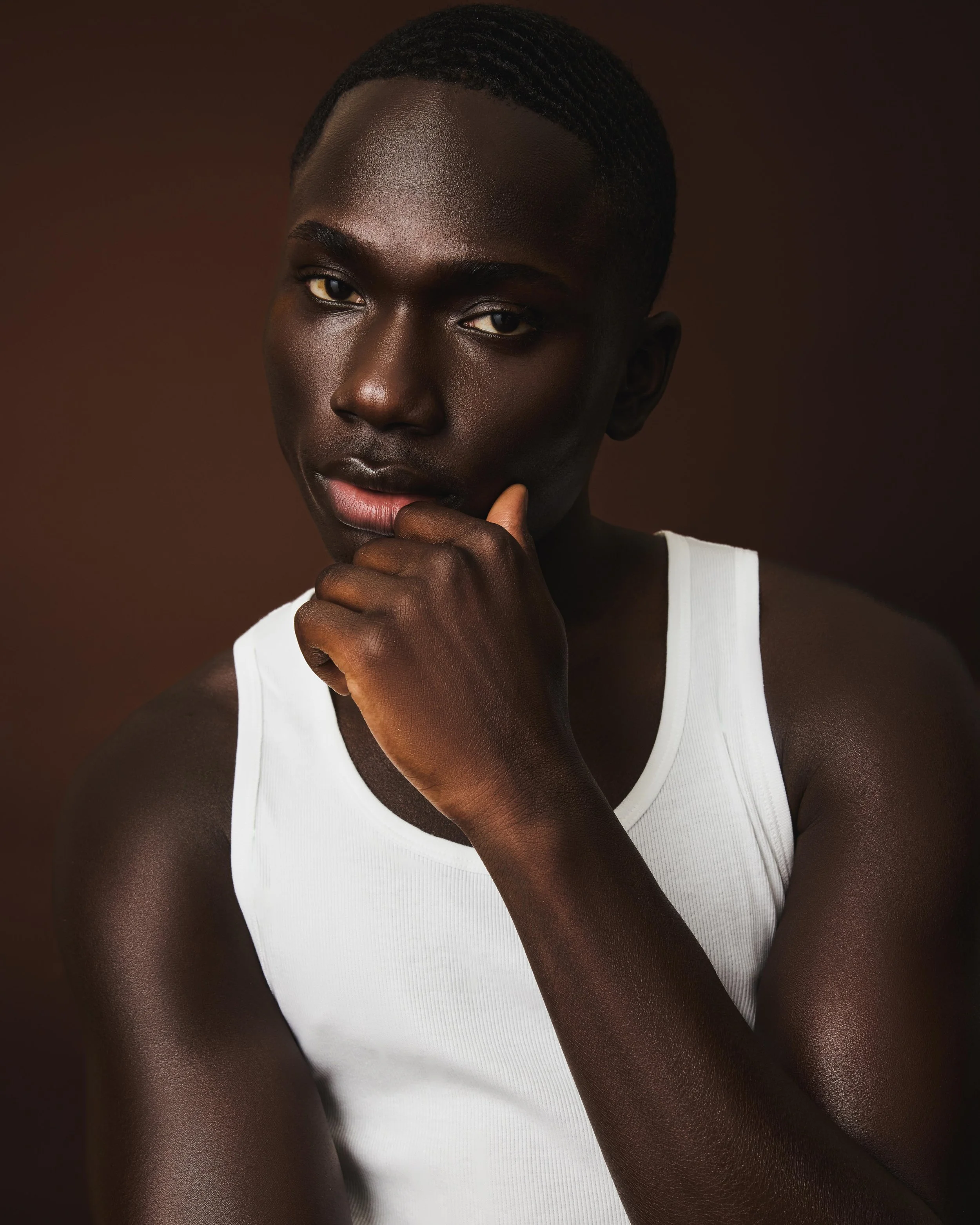 A close-up portrait of a young African American man with dark skin, short hair, wearing a white tank top, with thoughtful expression, touching his chin with his hand, against a plain, dark background.