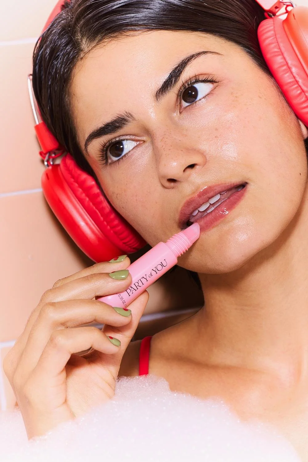 A young woman with dark hair, freckles, and makeup, wearing red headphones, is lying in a bubble bath and holding a pink lip balm labeled 'PARTY of YOU.' She is touching the lip balm to her lips and looking to the side with a relaxed expression.