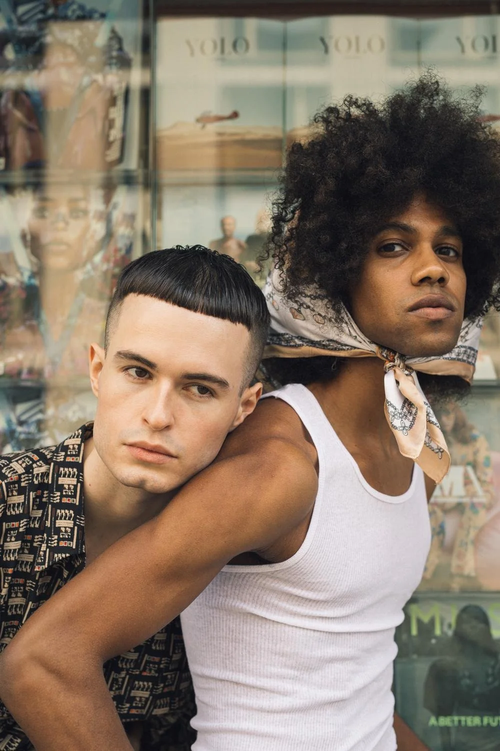 Two young men posing closely in front of a store window displaying dolls and shoes, one with short black hair and the other with curly black hair and a patterned headscarf, both looking at the camera.