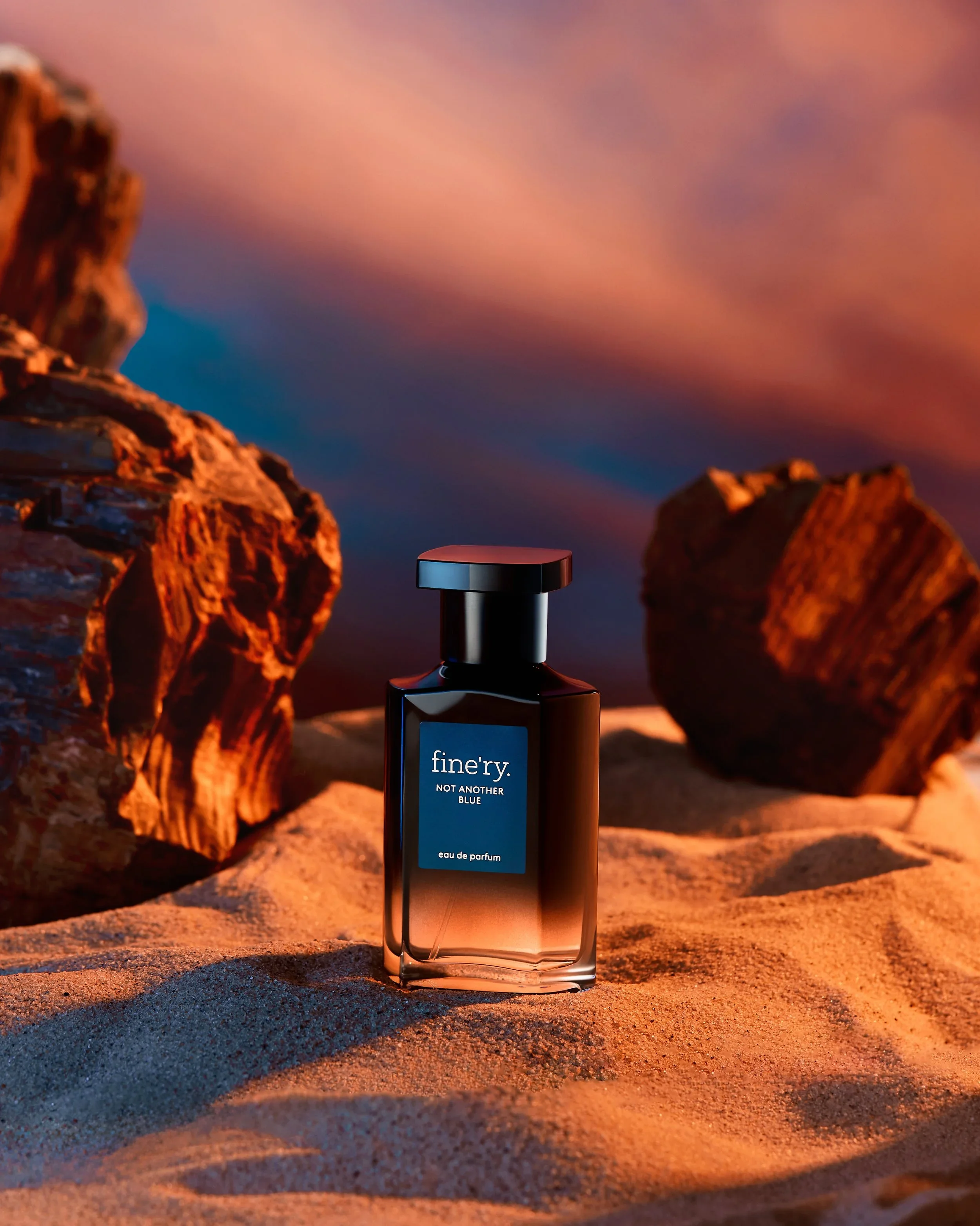 A perfume bottle labeled 'fine'r. Not Another Blue' sitting on sandy ground at sunset with large rocks and a colorful sky in the background.