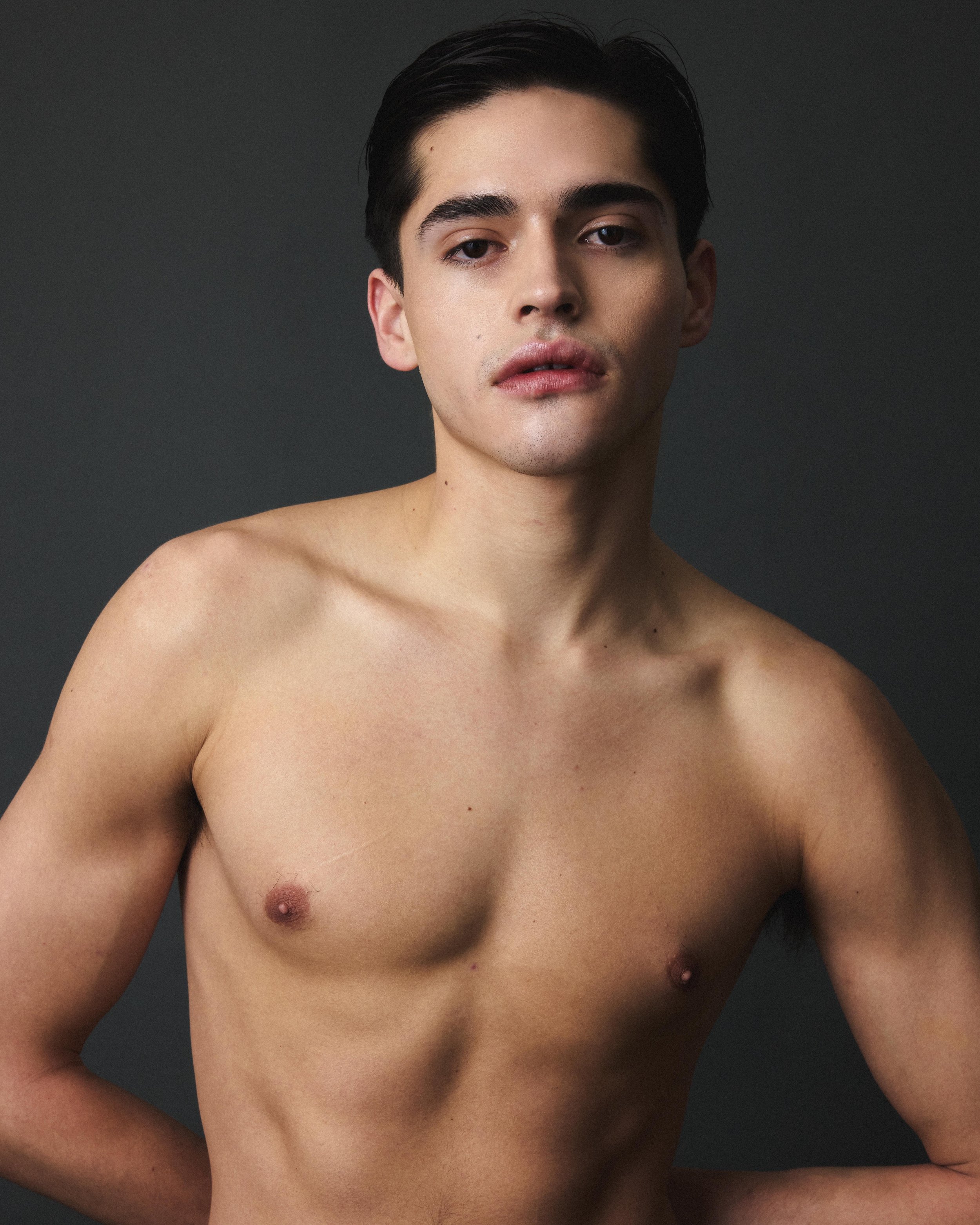 Portrait of a young man with short dark hair and light skin, looking directly at the camera, against a plain dark background. He is topless, showing his shoulders, chest, and arms.