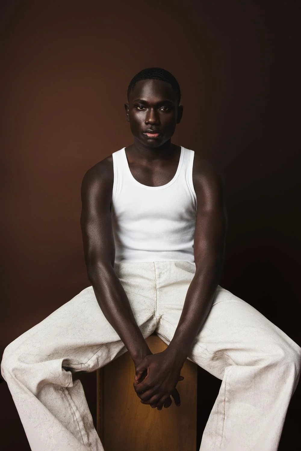 A young Black man wearing a white tank top and beige pants sitting against a dark brown background.