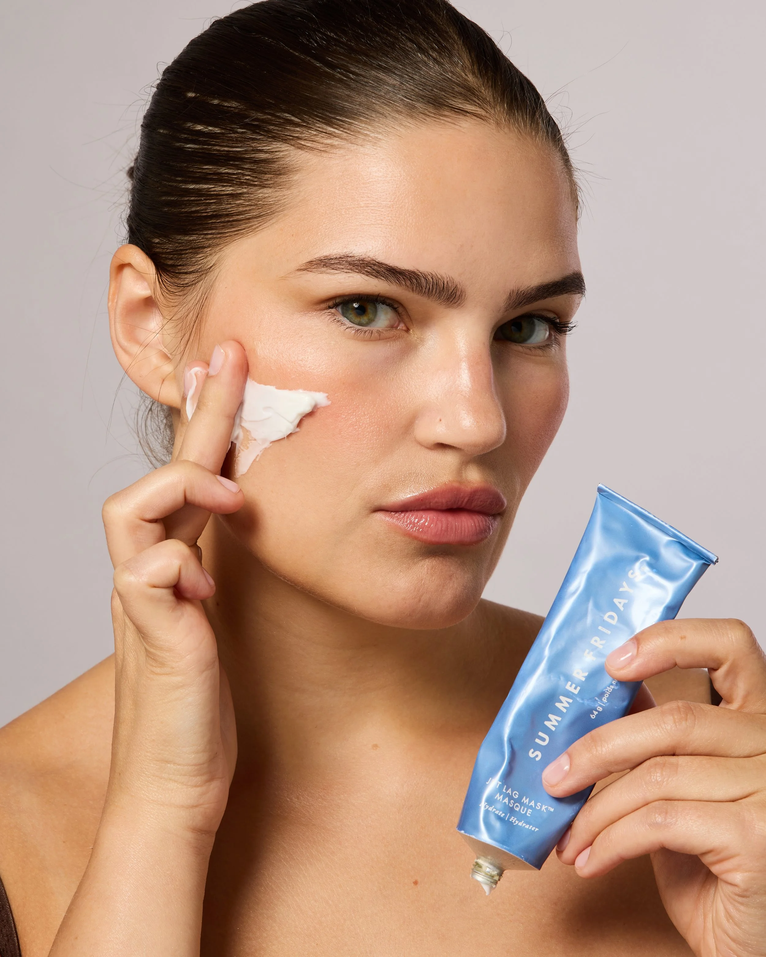A woman with green eyes and dark brown hair applying a white skincare mask to her cheek from a blue tube labeled 'SUMMER IDAL MASK'.