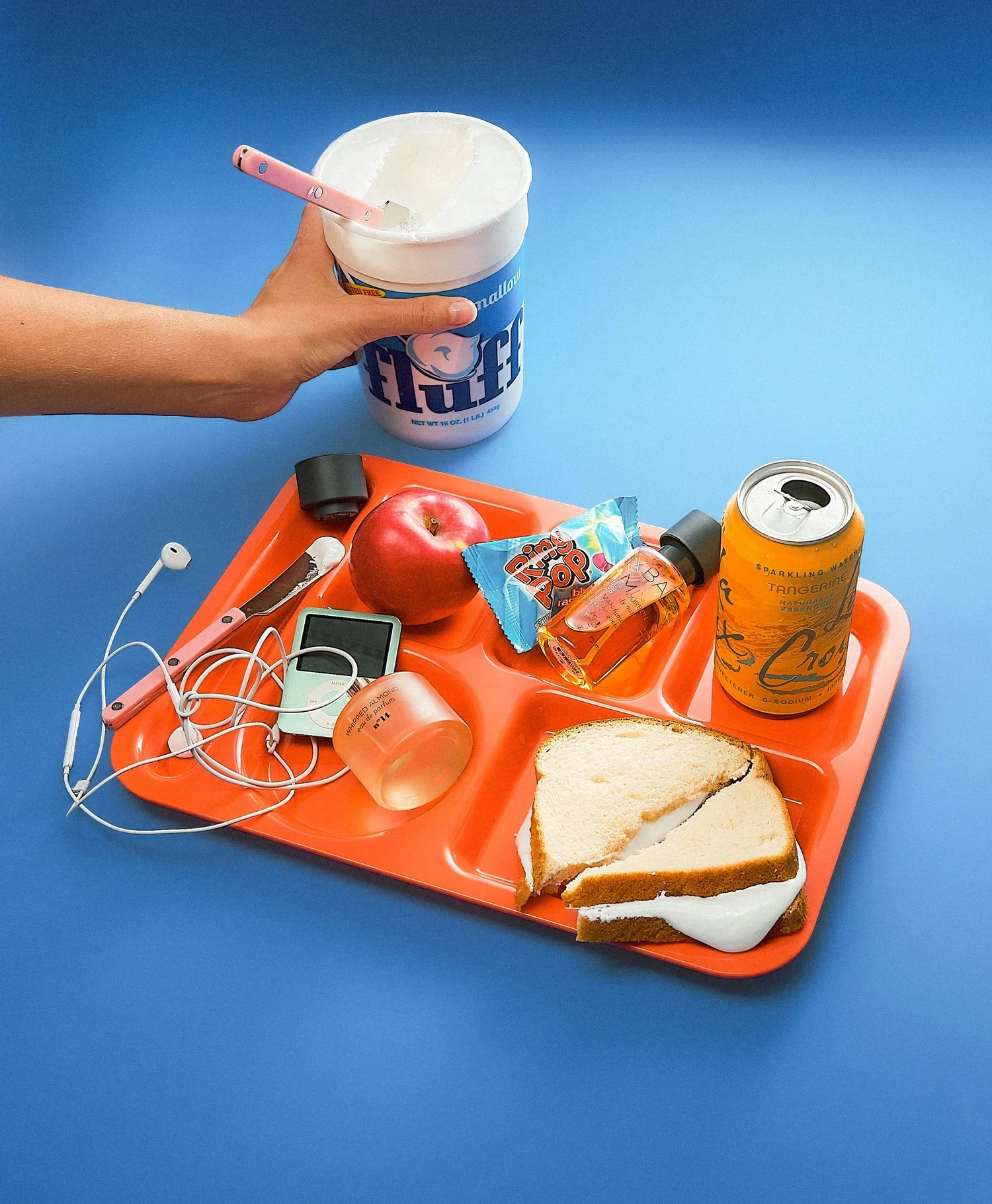 A blue table with an orange tray holding a piece of sliced bread with icing, a red apple, a packet of Ring Pop, an orange canned beverage, a small bottle of perfume, a digital timer, tangled white earphones, and a small digital device.
