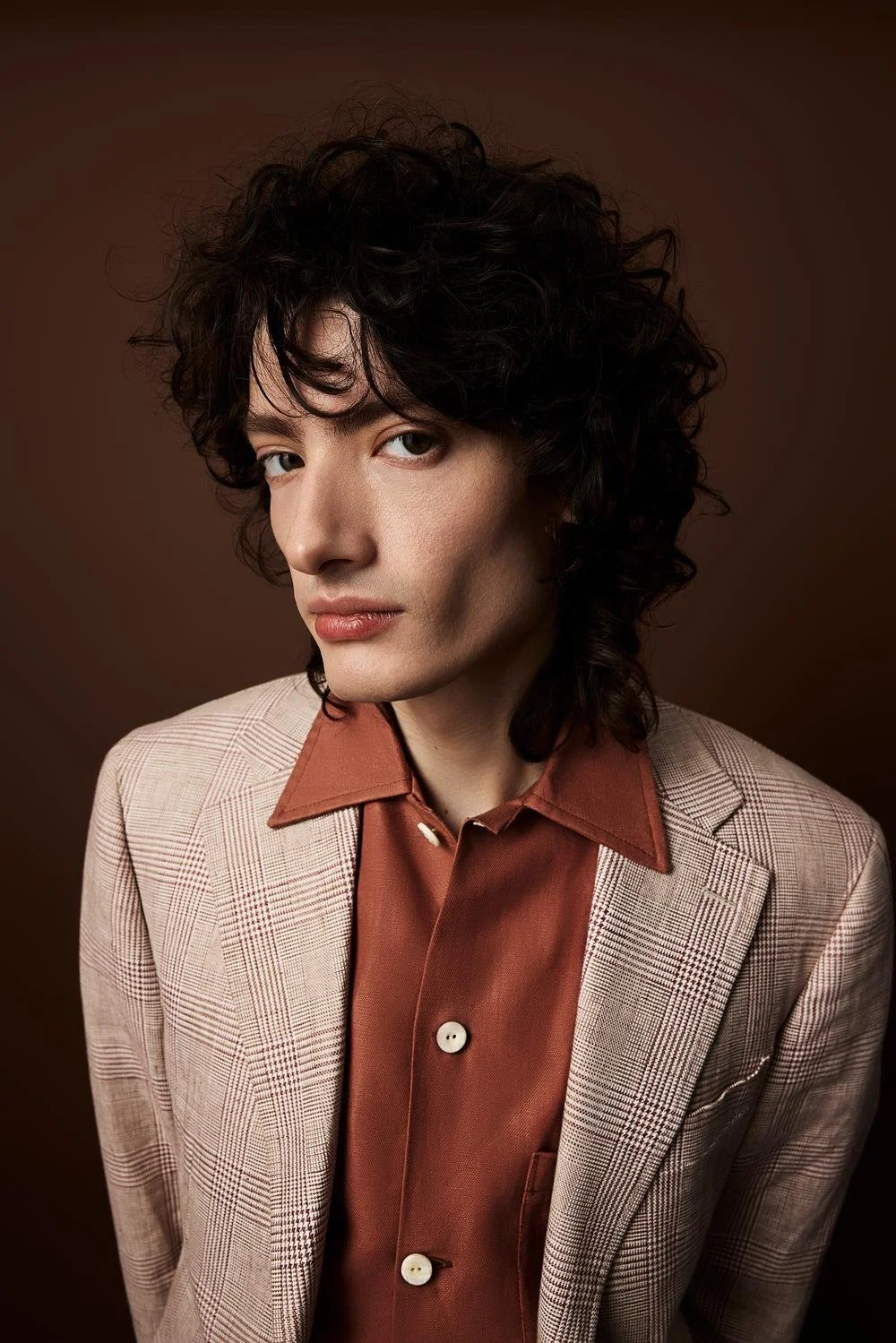 Portrait of a person with curly dark hair, wearing a beige checkered blazer and a rust-colored shirt, against a brown background.