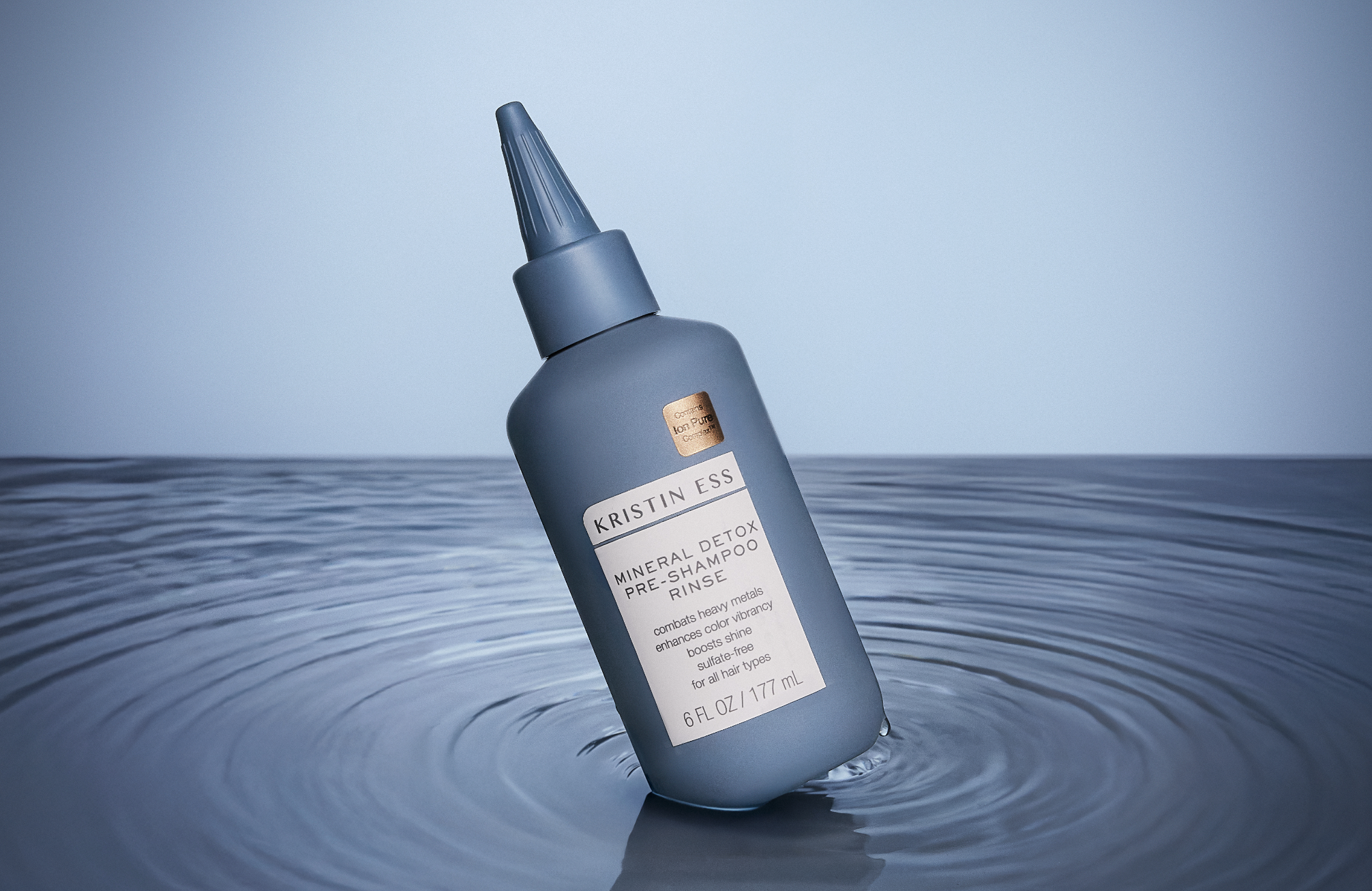 A bottle of Kristin Ess mineral detox pre-shampoo rinse floating on water with a light blue background.