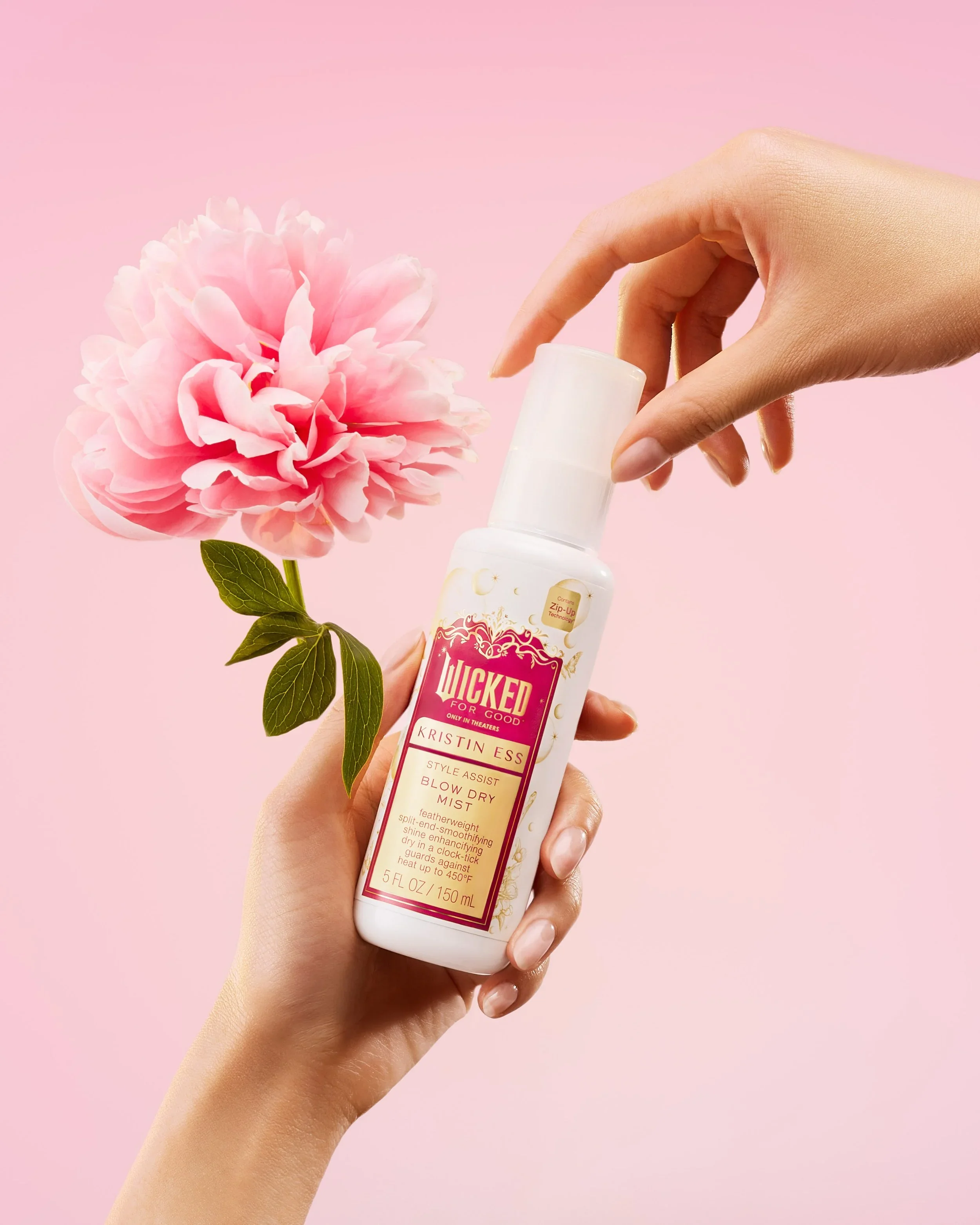 A person holding a bottle of Kristin Ess Style + Assist Blow Dry Mist with a pink flower and green leaves against a pink background.