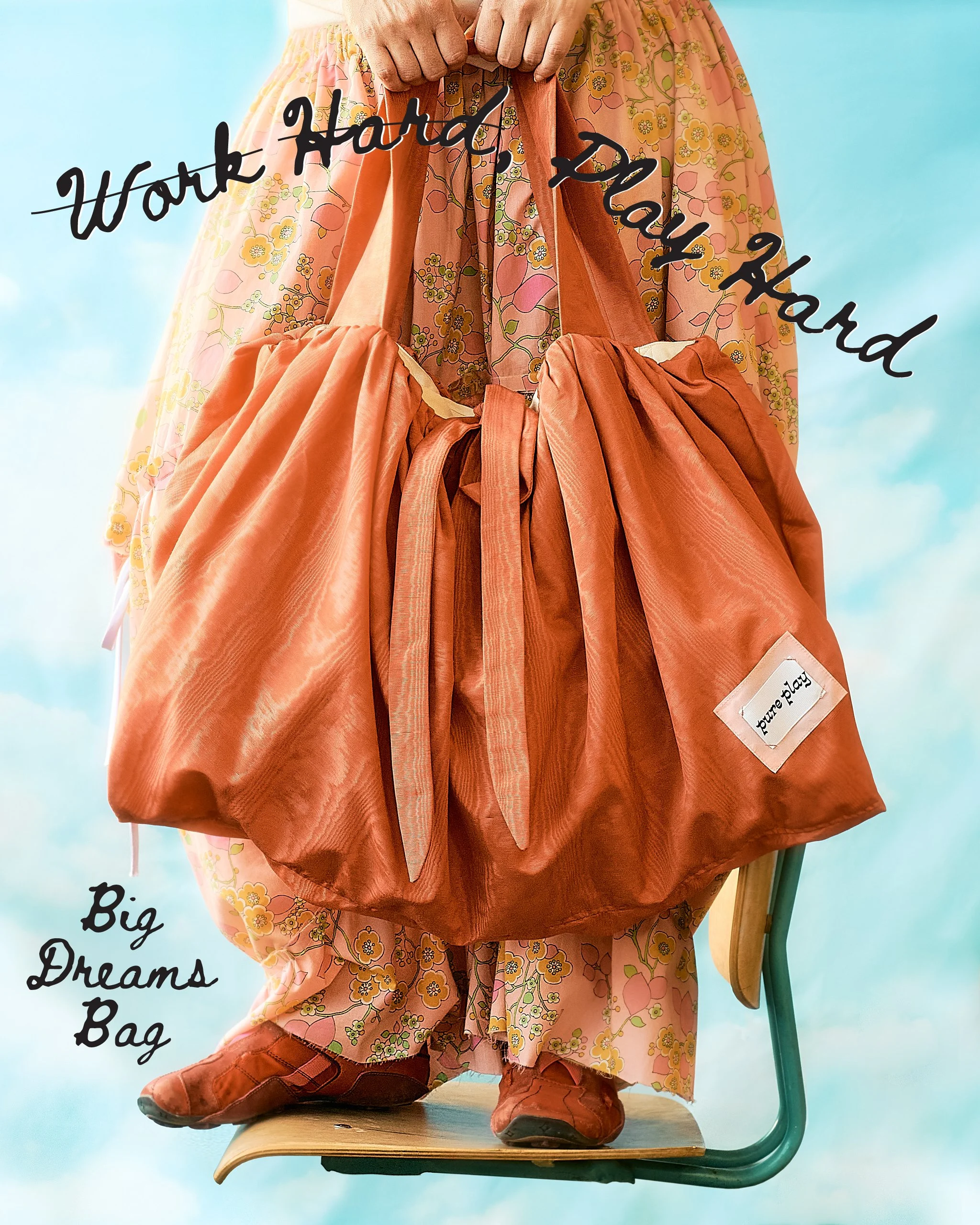 A person standing on a floating school chair with a sky background wears red shoes and pink floral pants holding a large red bag. The image has text overlay that reads, 'Big Dreams Bag' & "Work Hard, Play Hard", but, 'Work Hard' is crossed out.