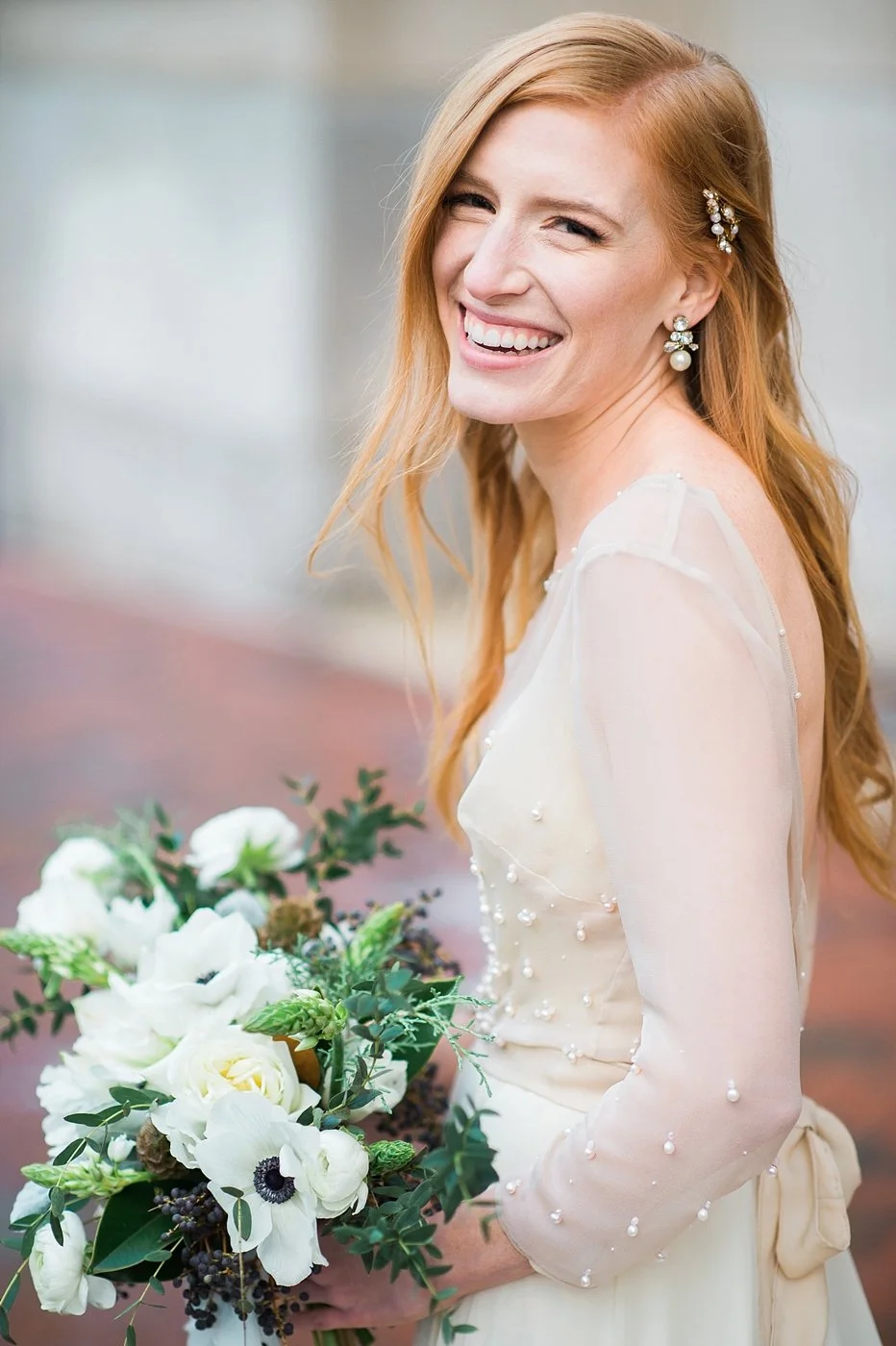 A smiling red-haired woman in a white dress with pearl accents, holding a bouquet of white and green flowers.