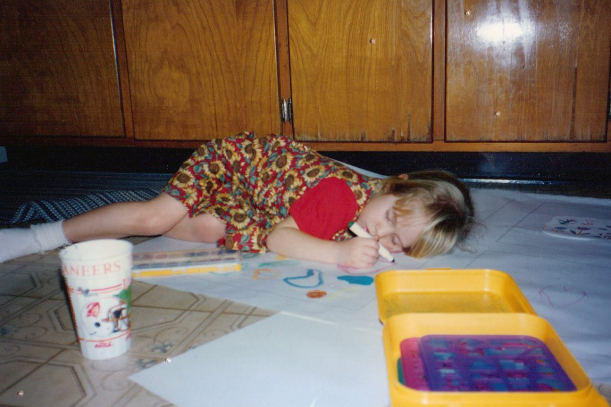 A young girl in a sunflower dress lying on her side on the floor of a kitchen asleep, drawing on a large sheet of paper with markers. There's a yellow craft box and purple stencils nearby. The background shows wooden cabinets.