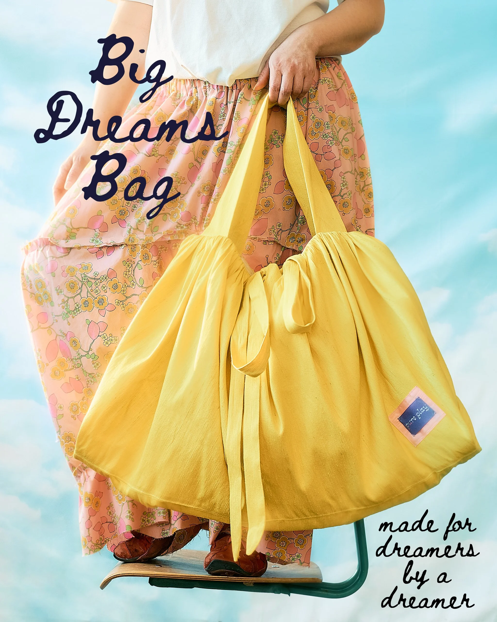 A person standing on a floating school chair with a sky background wears red shoes and pink floral pants holding a large yellow bag. The image has text overlay that reads, 'Big Dreams Bag' and part of another phrase, 'made for dreamers by a dreamer.'