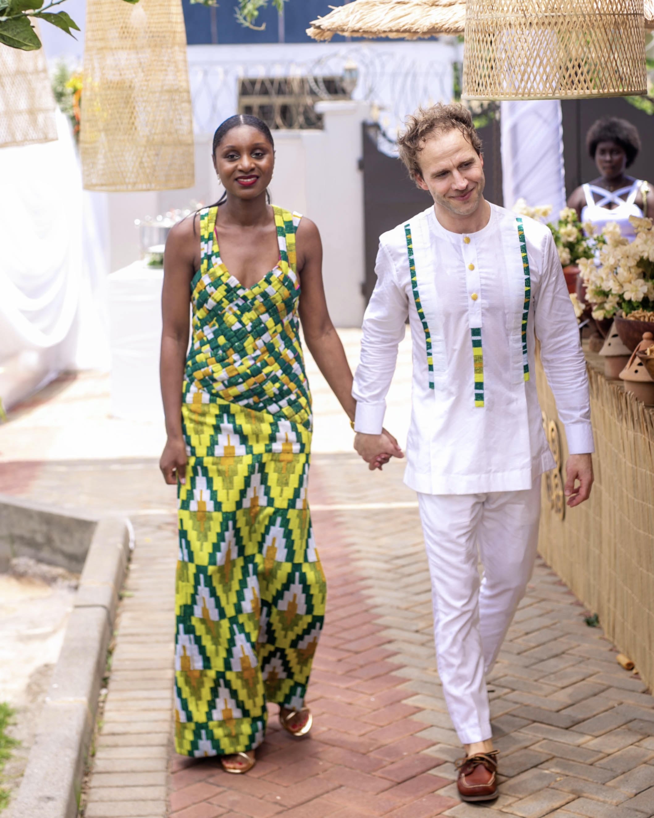 A man and a woman holding hands and smiling while walking outdoors on a brick pathway, with the woman wearing a colorful dress with a geometric pattern and the man wearing a white shirt with colorful accents.