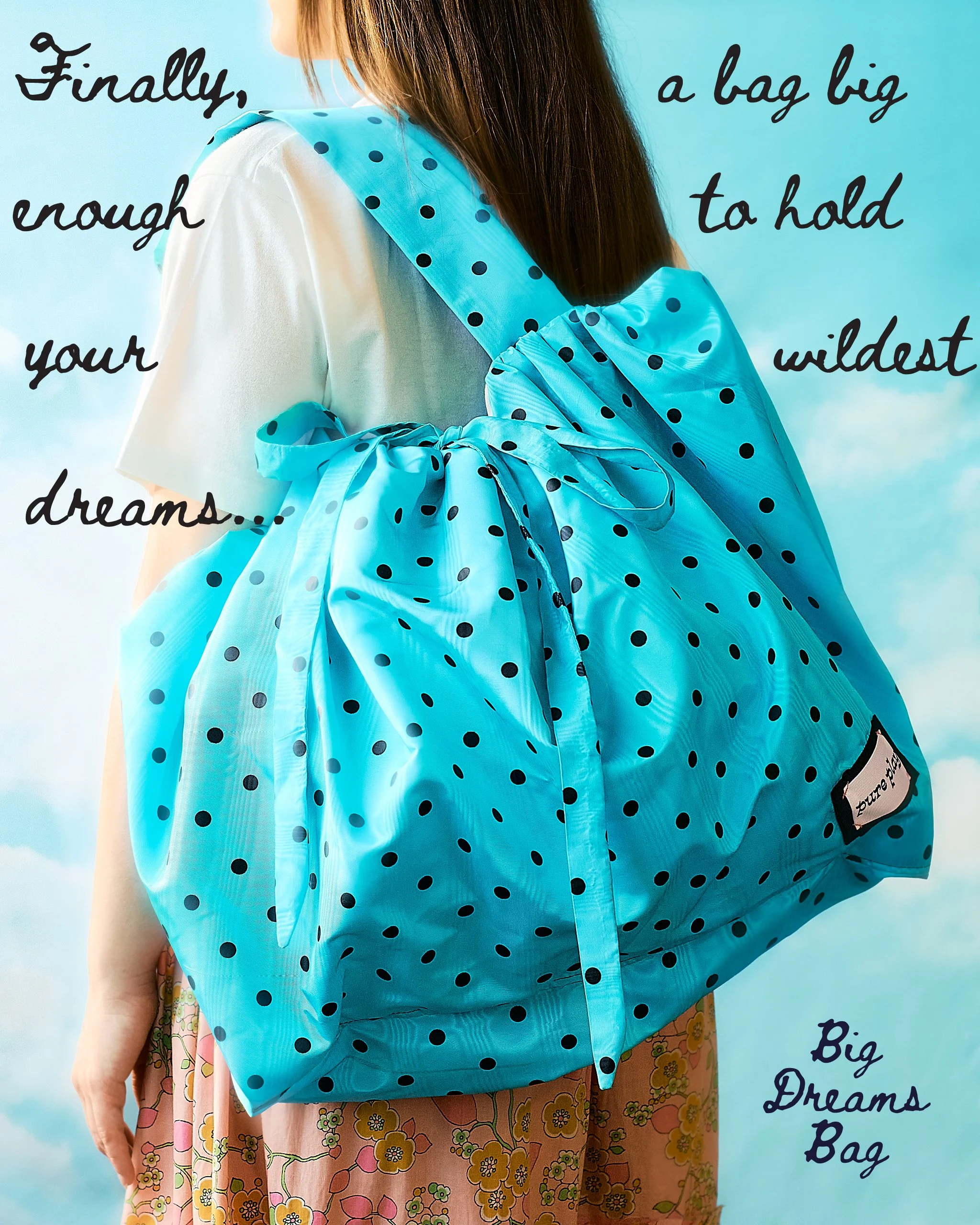 A person wearing a white shirt & floral skirt is carrying a large, teal polka dot bag over their shoulder with a sky background. The image has text overlay that reads, 'Big Dreams Bag' & "Finally, A Bag Big Enough to Hold Your Wildest Dreams."