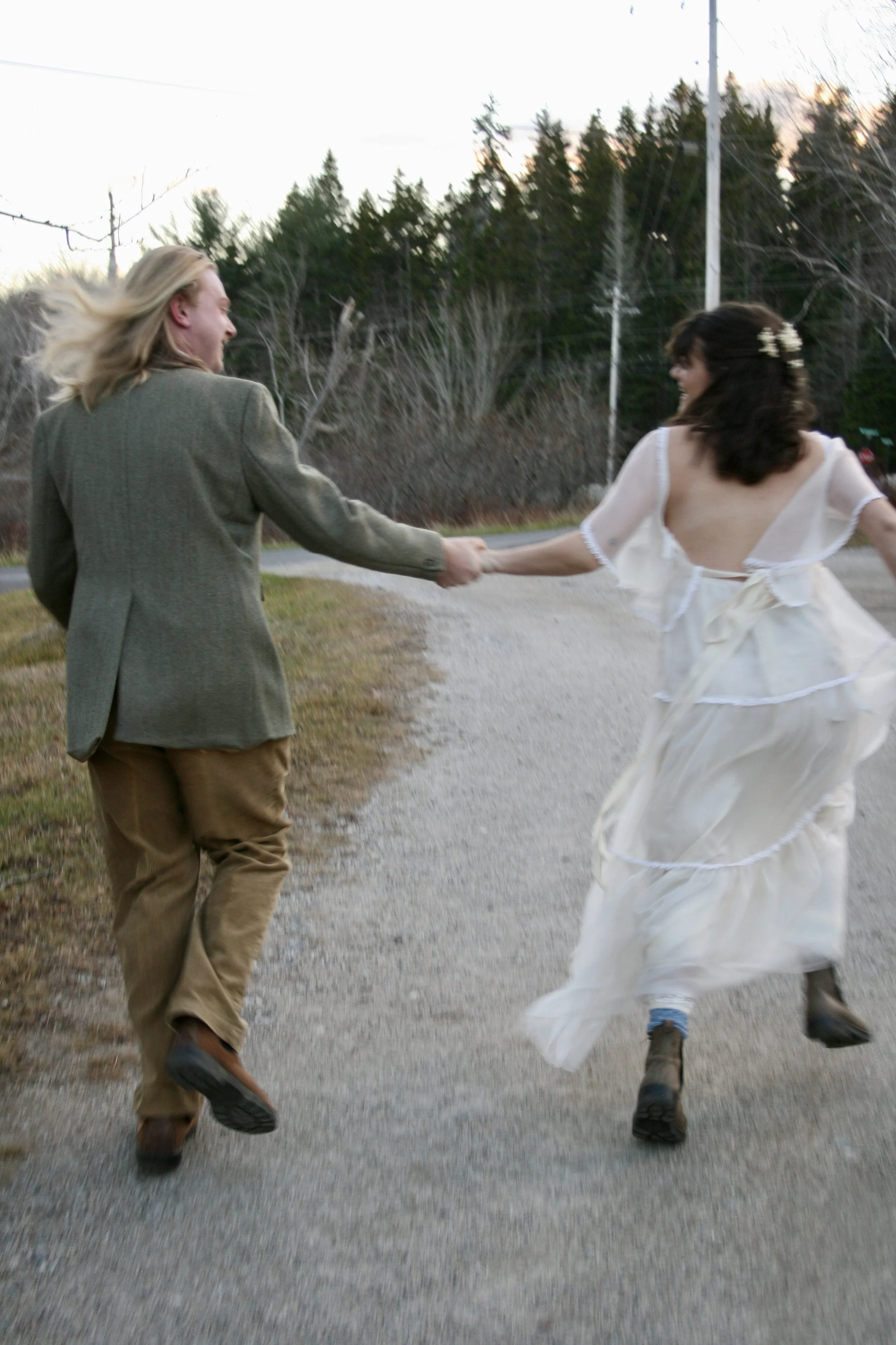 Two people, a man and a woman, are holding hands and running on a rural road. The man is dressed in a blazer and khakis, and the woman is wearing a white dress with lace details. There are trees and utility poles in the background, and the sky appears to be at sunset.