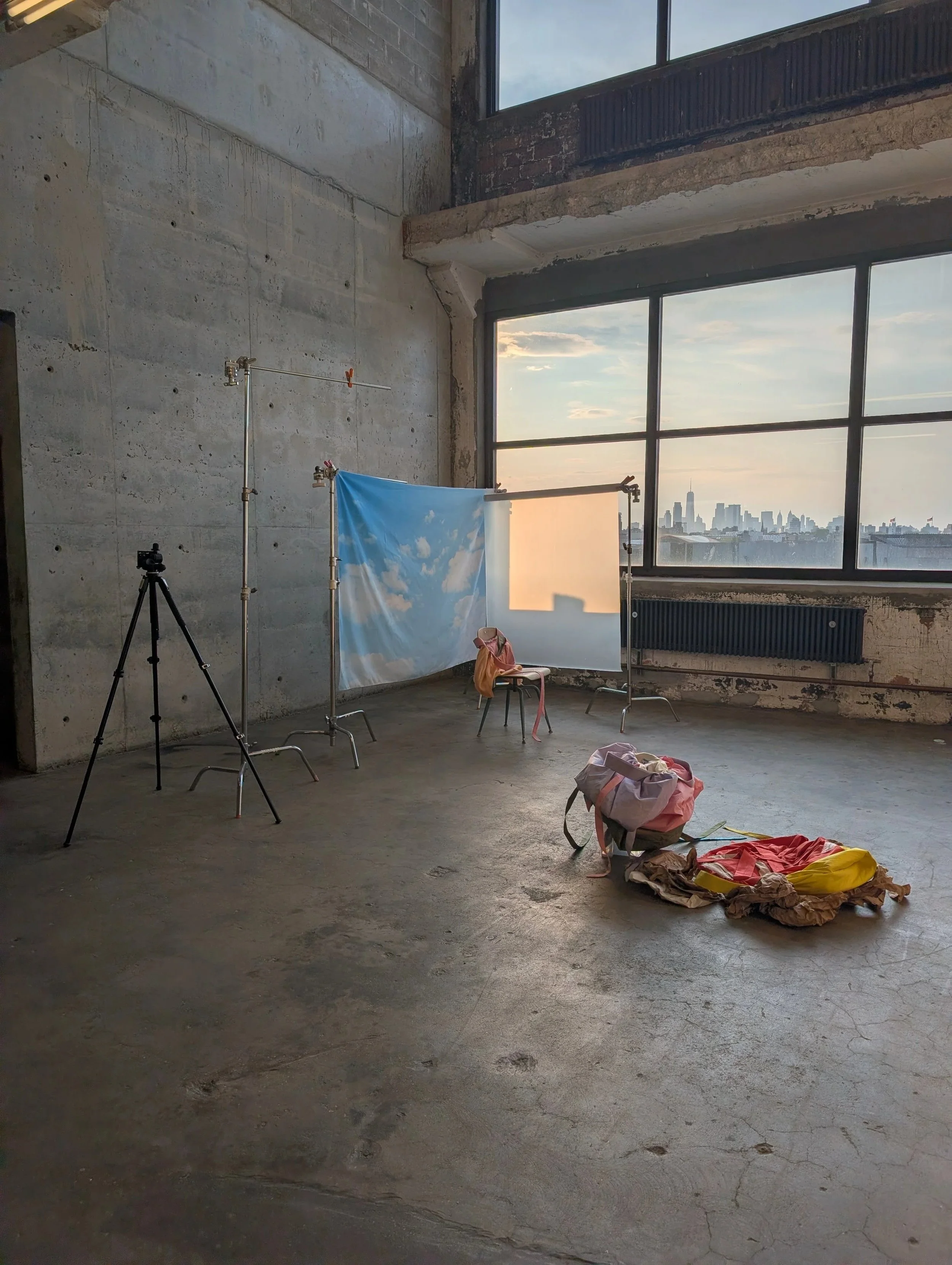 An unfinished industrial-style room with large windows, exposed brick and concrete walls, and miscellaneous items including a camera on a tripod, a backdrop with sky and clouds, a pink backpack, and some fabric on the floor.