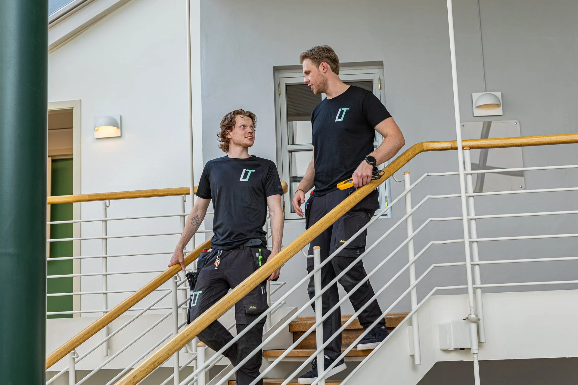 To menn har arbeidsklær med Lutech Elektro logo, snakker mens de går ned en trapp.