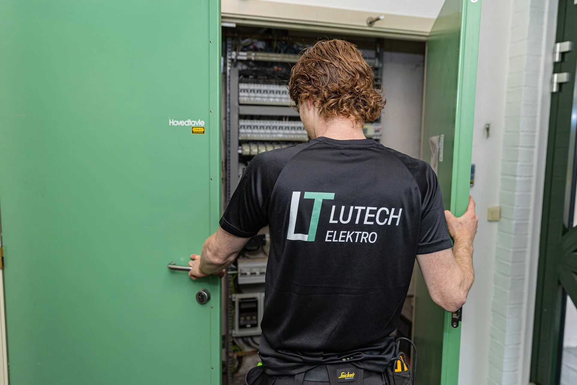 En person står foran en åpen elektrisk skap, iført en svart T-skjorte med teksten 'Lutech Elektro' på ryggen. Han jobber med elektriske ledninger.
