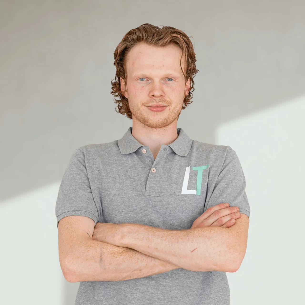 En ung mann med krøllete hår, kledd i en grå polo, står med armene i kryss mot en lys bakgrunn.