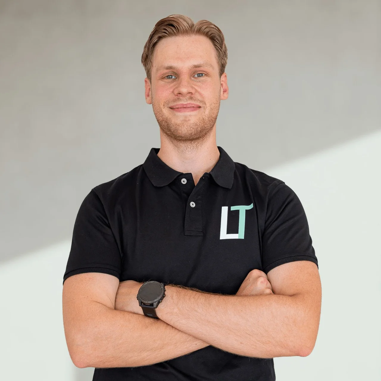En mann med blondt hår, kledd i svart polo med logo 'LT' på brystet, står med armene krysset foran en lys, minimalistisk vegg.