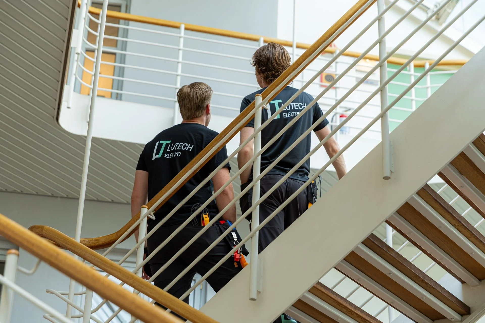 To arbeidere som jobber på en trappestige i en innendørs setting, kledd i arbeidsklær med logoen "Lutech Elektro" på ryggen, omgitt av en moderne bygning med trapper og gelender.