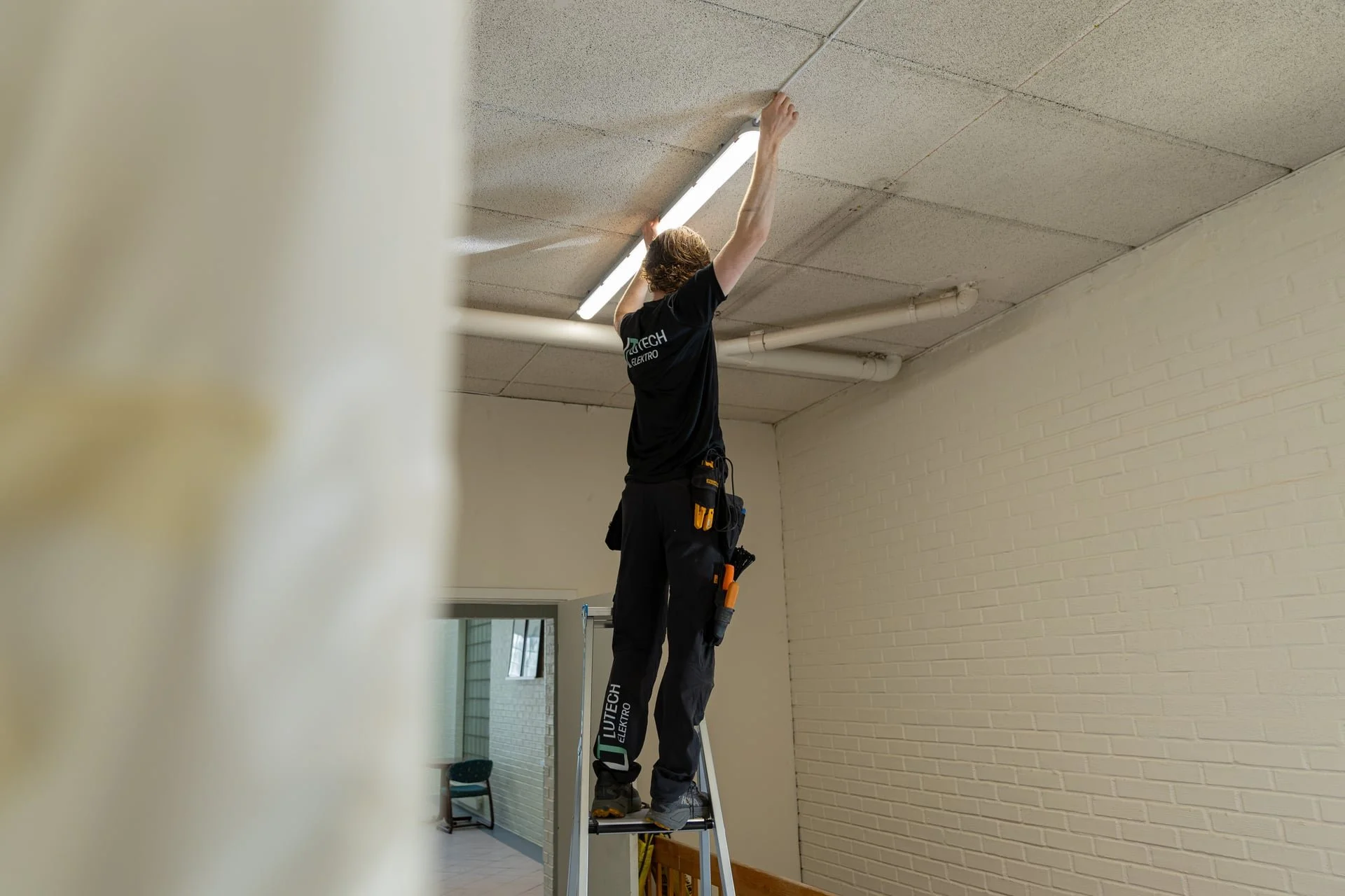 Person som står på en oppstigningsluke på en stige, som jobber med å installere eller vedlikeholde en lysarmatur i taket.