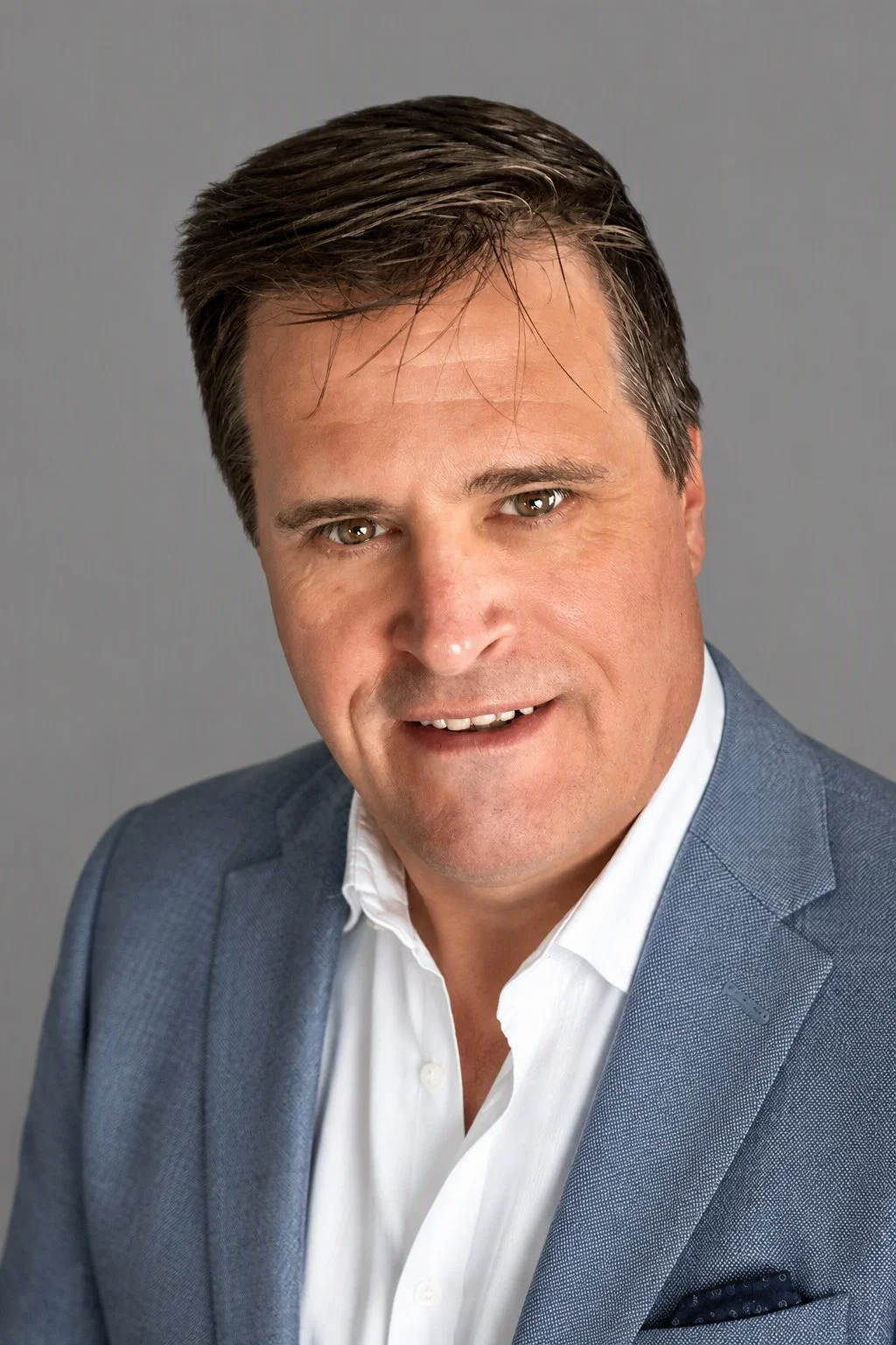 Headshot of a middle-aged man with dark hair wearing a light blue blazer and white dress shirt against a neutral background.