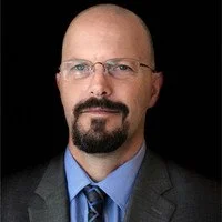 A man with glasses and a goatee wearing a suit and tie, posing against a black background.