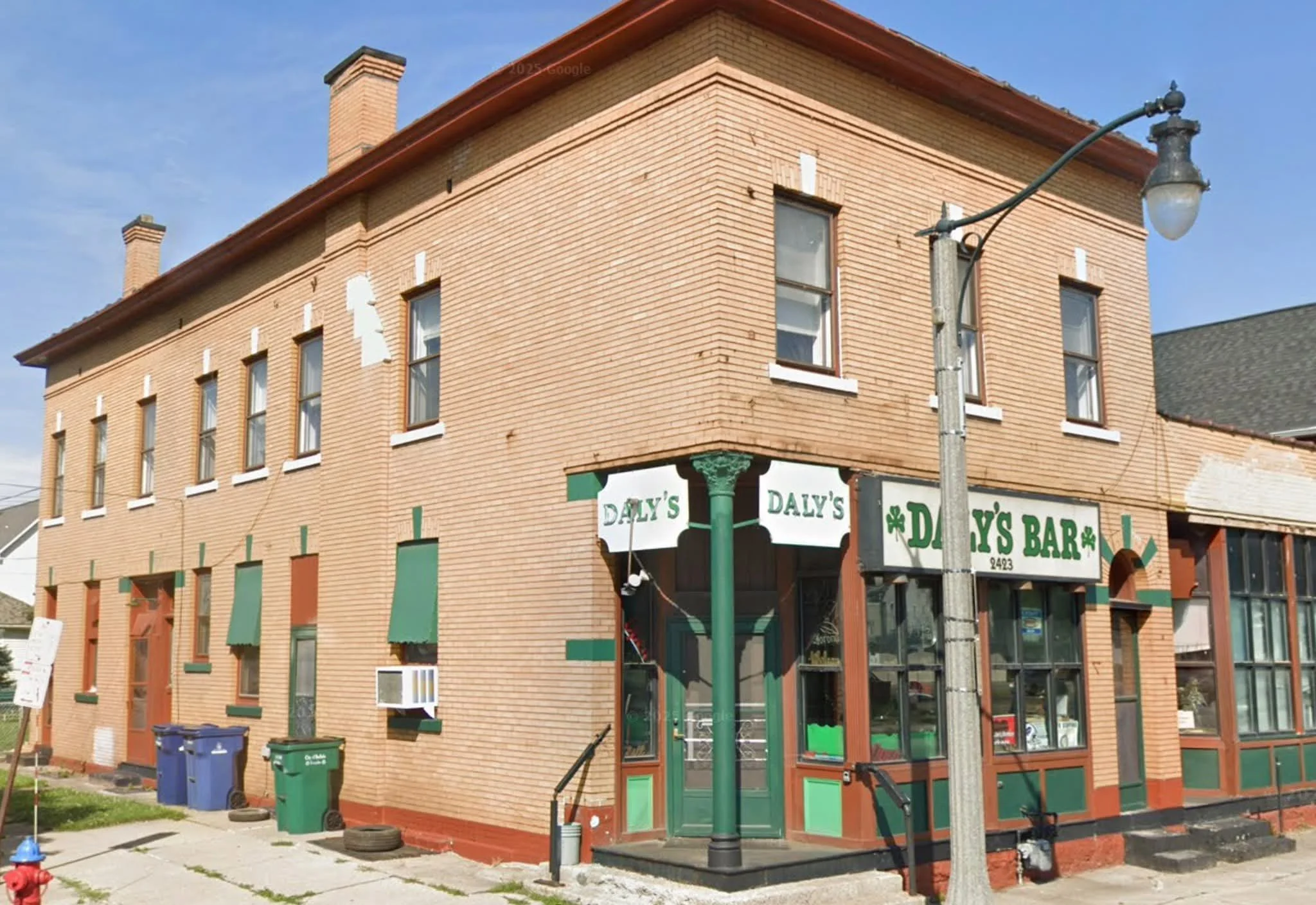 Corner view of a brick building with commercial signage for Daly's Bar, green door and window trims, and a sidewalk with garbage bins in front. A streetlamp is visible on the sidewalk, and the sky is clear with a few clouds.