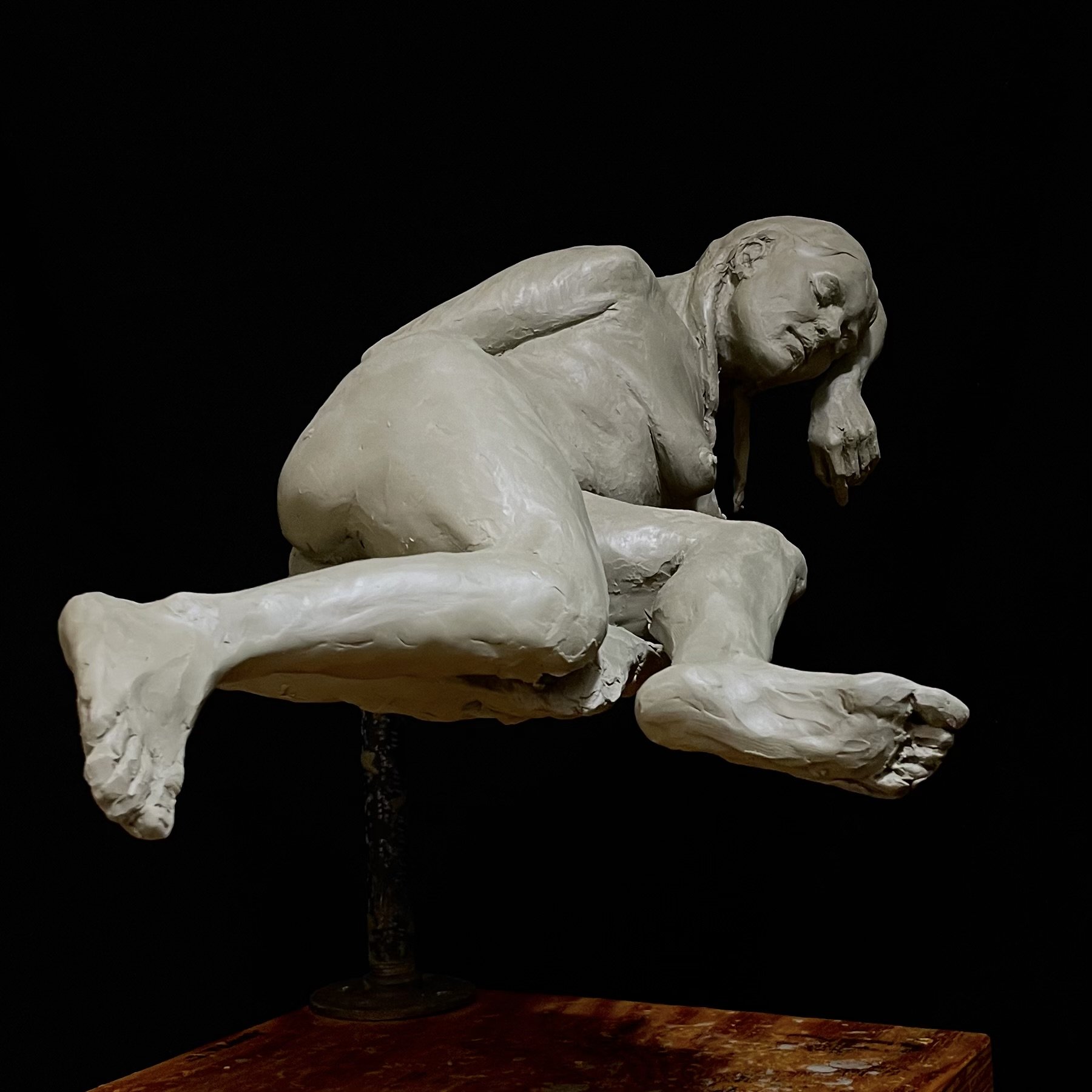 A clay sculpture of a woman reclining with her head resting on her hand, displayed against a black background.