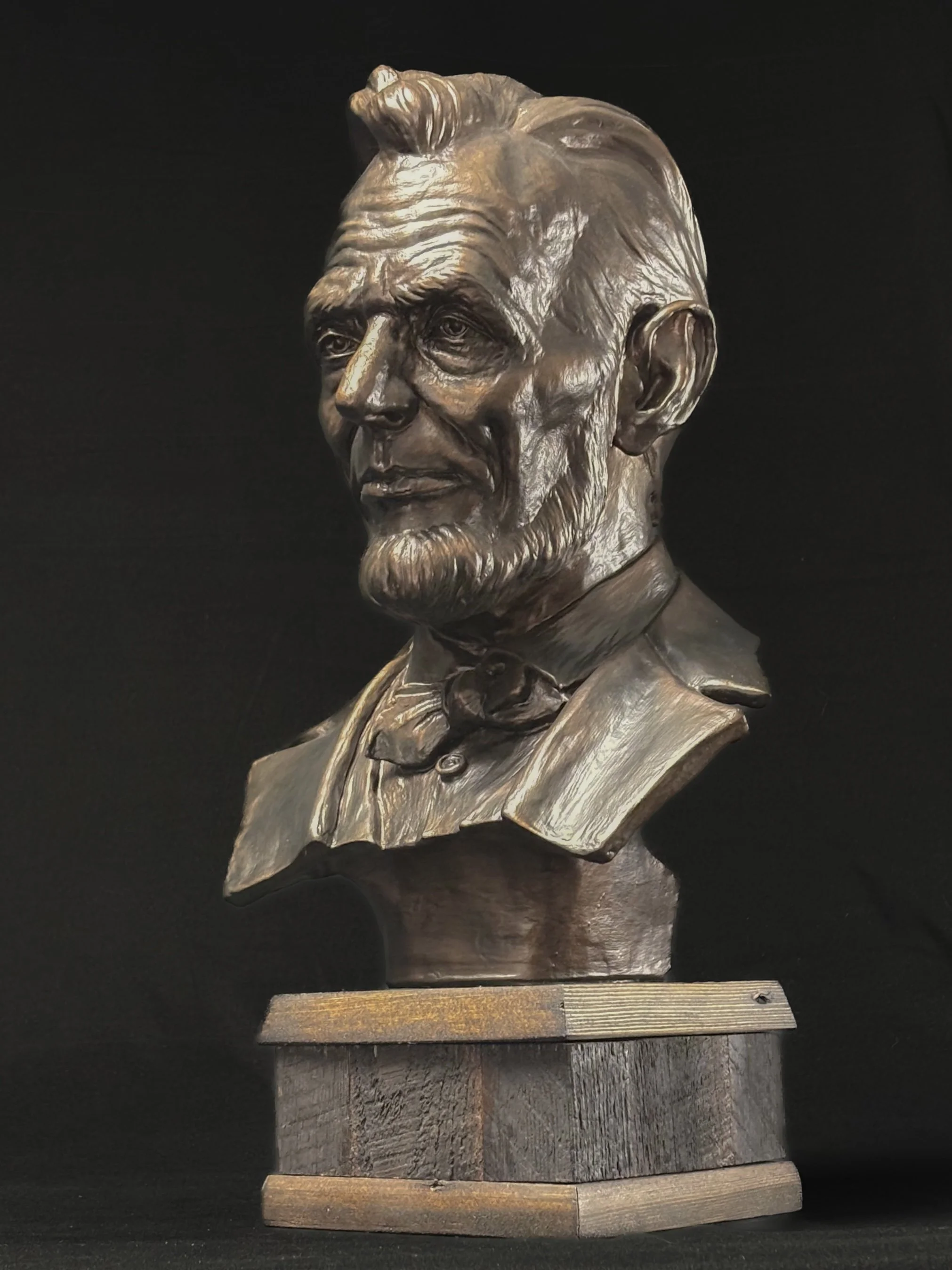 Bronze bust sculpture of Abraham Lincoln, depicted with a serious expression, neatly combed hair, and dressed in a suit with a bow tie, mounted on a wooden base.