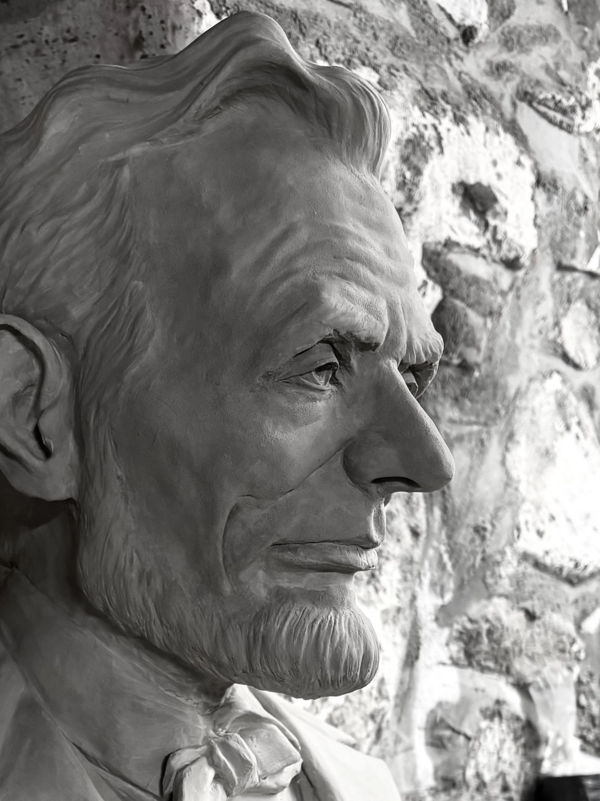 Close-up of Abraham Lincoln's bust sculpture showing his profile with detailed facial features, beard, and hairstyle, set against a stone wall background.