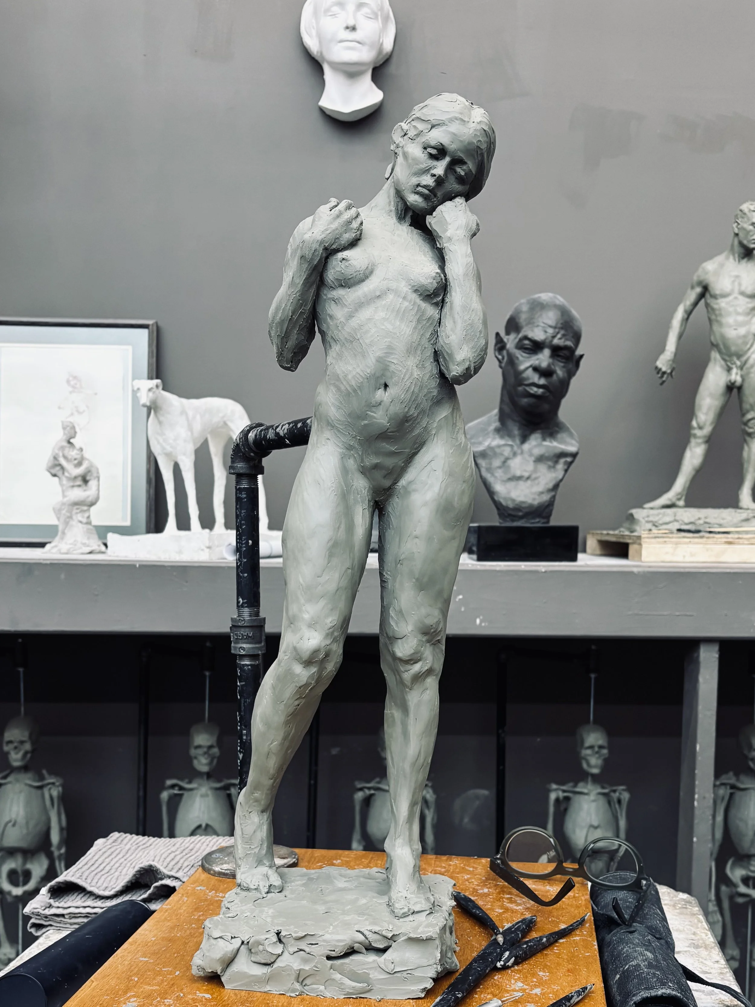 Clay sculpture of a woman with fists clenched and head slightly tilted, on a worktable surrounded by sculpting tools, with other sculptures and head busts displayed in the background.