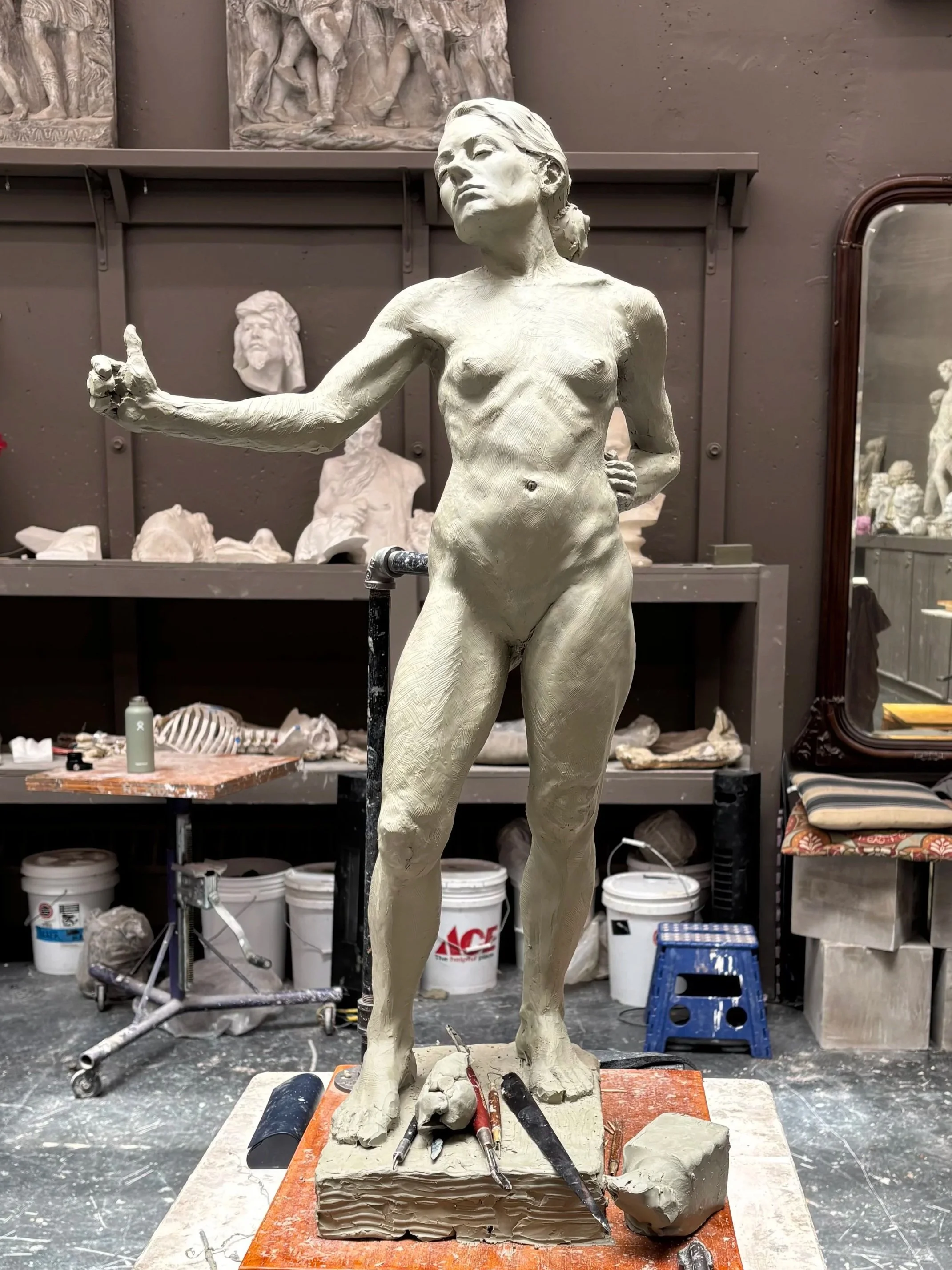 A clay sculpture of a nude woman standing on a work table in an artist's studio, with art supplies and sketches in the background.